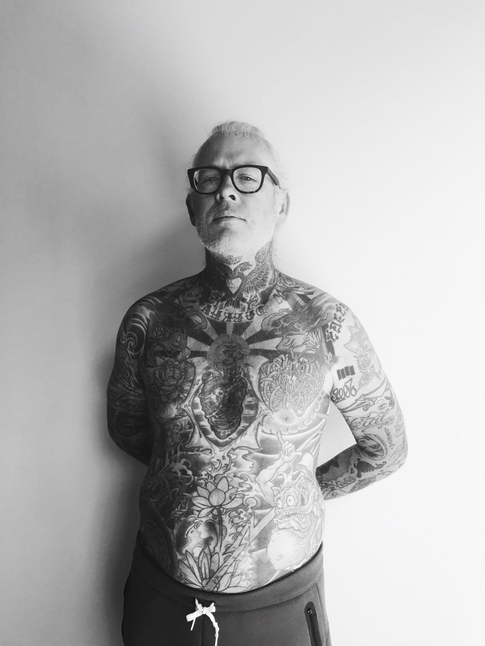 Black and white portrait of mike giant in front of a white wall. He is wearing black galsses. He is shirtless and his torso is covered in tattoos. 