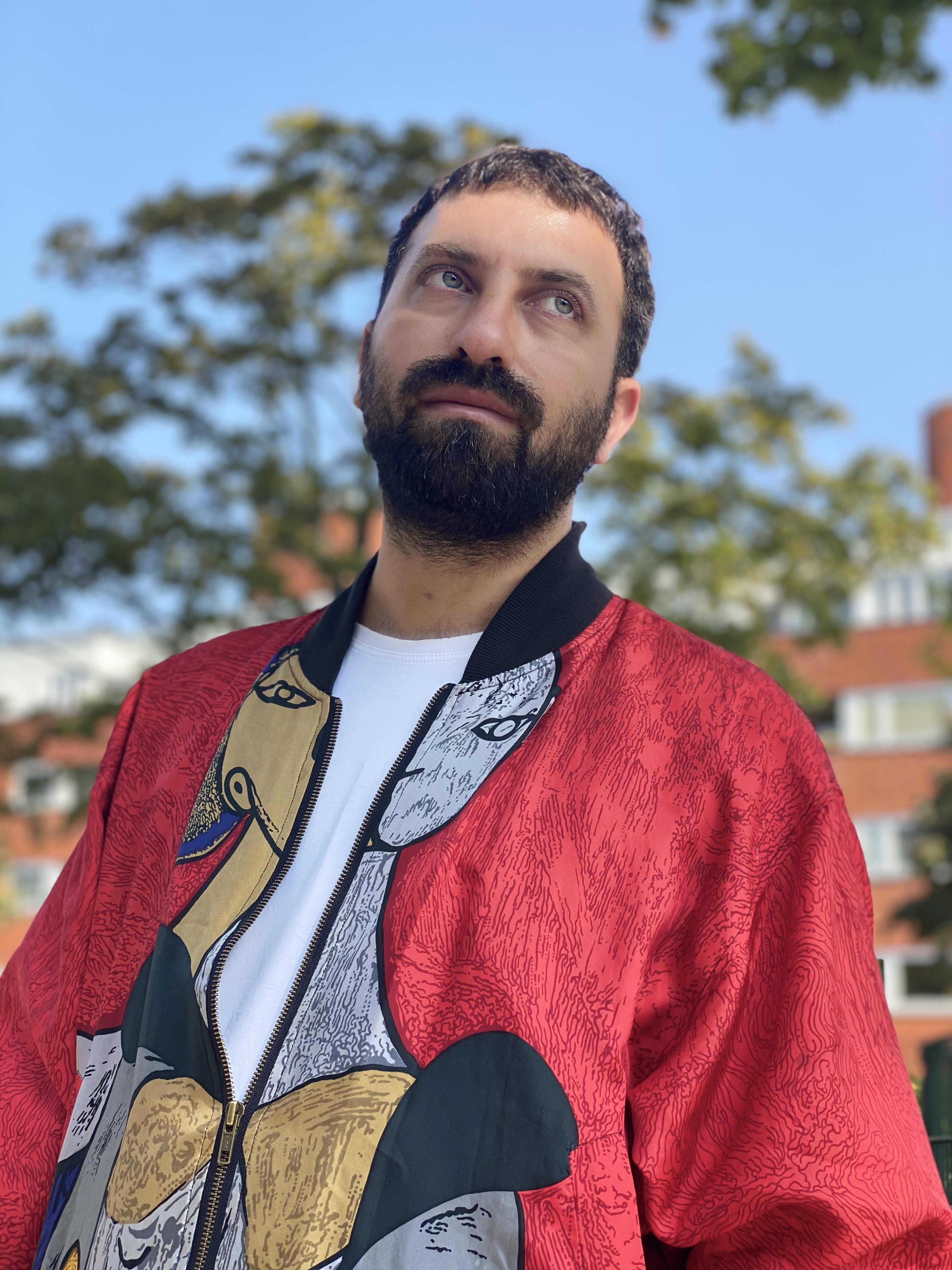 A portrait of Ahmet against a blue sky and a few large trees. He is looking off behind the camera, and is wearing red bomber jacket with a design on it over a white tee. He has a thick beard and a serious look on his face. 