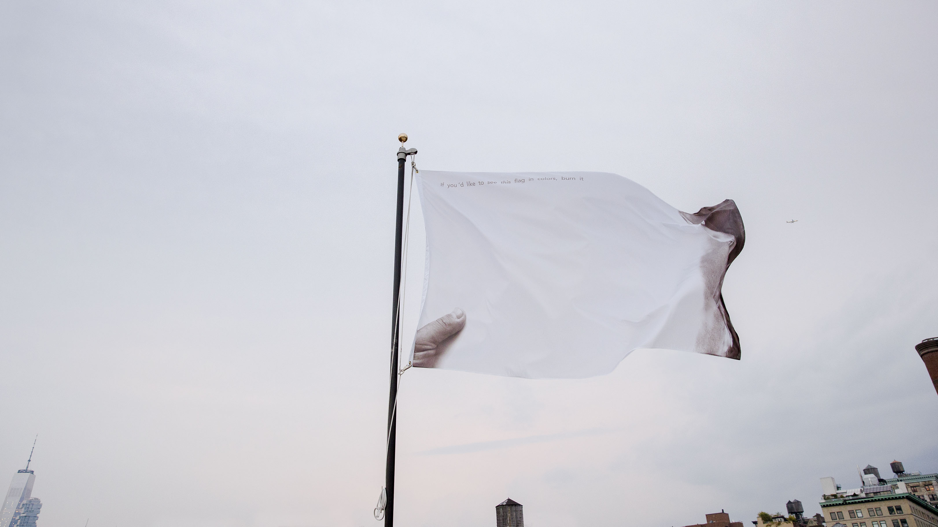 A white flag flying in a gray sky. The flag has a thumb illustrated in the bottom left corner. The small text spans across the top of the flag and reads: “If you’d like to see this flag in colors, burn it.”