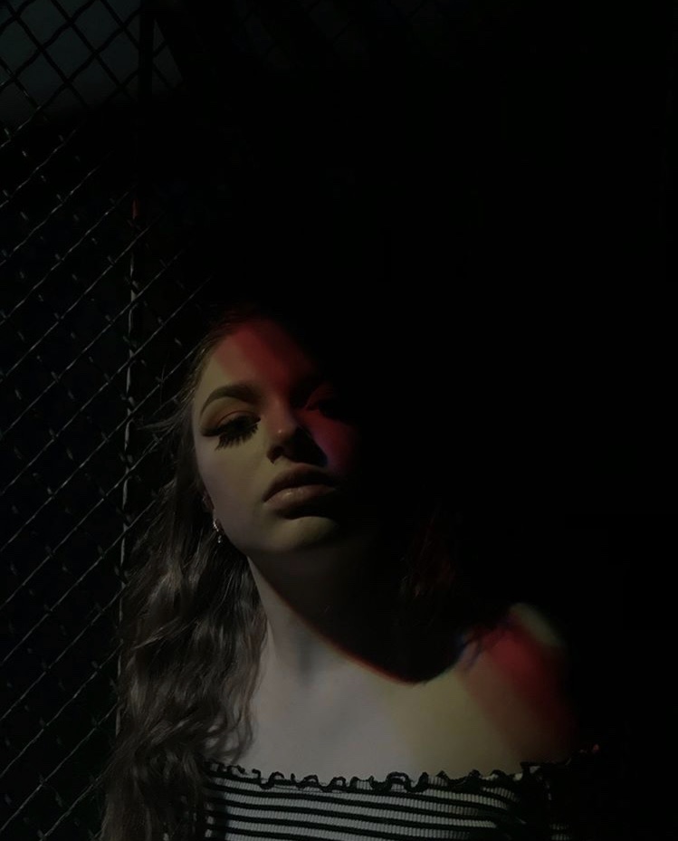 A young woman leans against a chain link fence at night. Half her face is shrouded by shadows and she looks neutrally into the camera. 