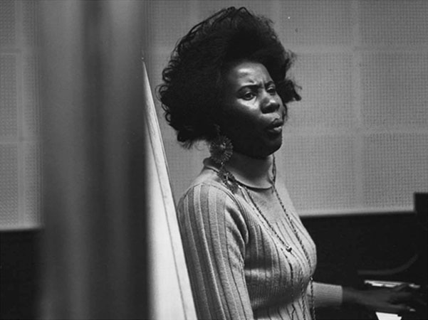 black and white photo of Alice Coltrane sitting at a piano, looking off to the right