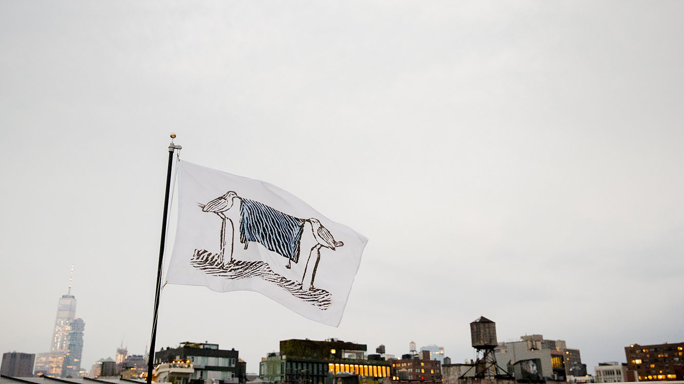 White flag flying against an urban landscape. On the flag is a drawing of two birds standing on a post holding a blue and black stripped cloth in their beaks.