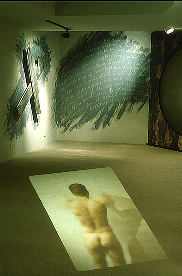Photo of mixed media installation. a projected photo of a naked man from behind. On the walls is green paint with words etched in that are not legible and on the opposite wall, a wooden cross painted and hung at an angle