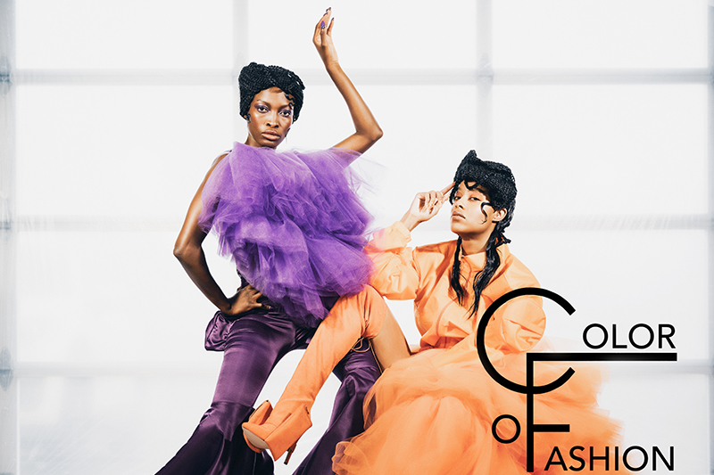 two black women in the center of the frame wearing couture dresses. One in purple and one that is orange with Color of Fashion logo in the bottom right corner