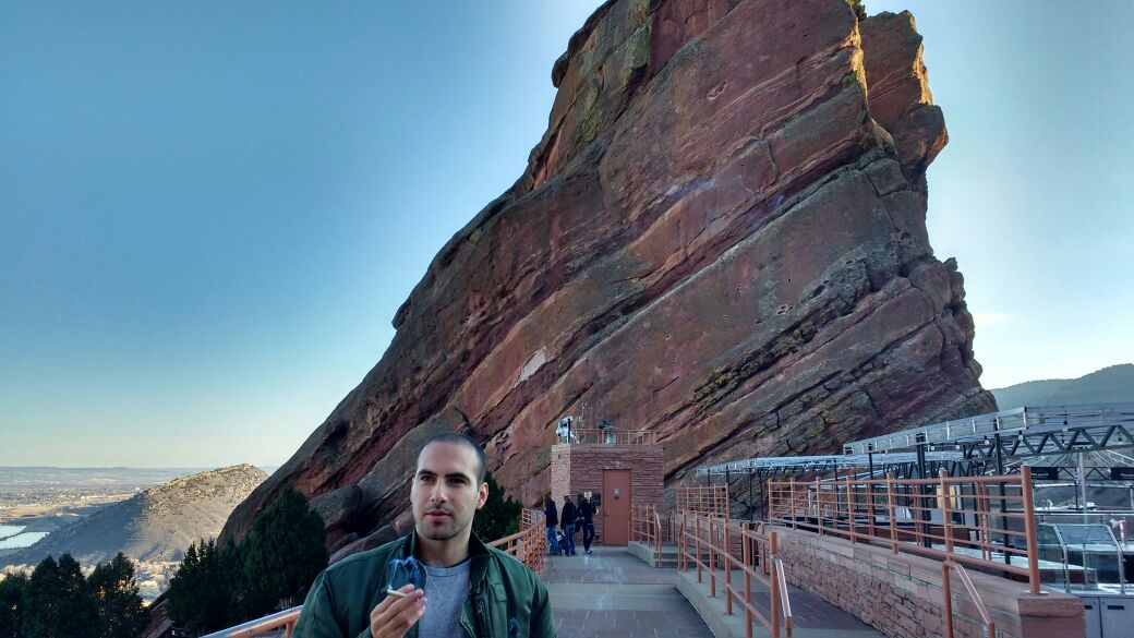 A man stands in front of red rocks. He is wearing a green jacket and holds a joint in between his fingers. 