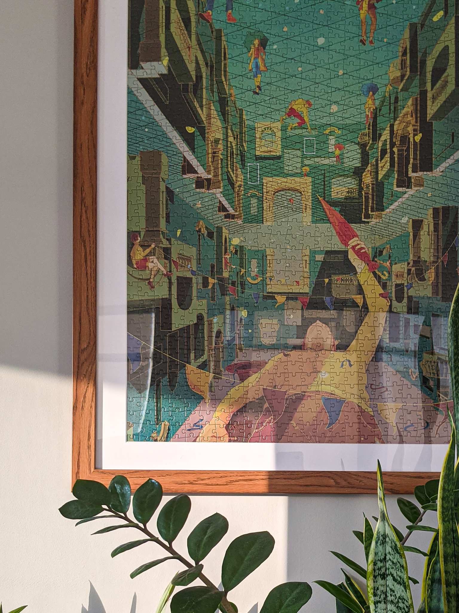 A completed puzzle in a brown frame and hung up on the wall. Light hits the bottom of the puzzle and plants take up the space underneath the puzzle. The image on the puzzle is a transcendent digital painting that looks like a structural study with characters throughout. The characters are on different levels of the scene and are holding closed and open umbrellas. 