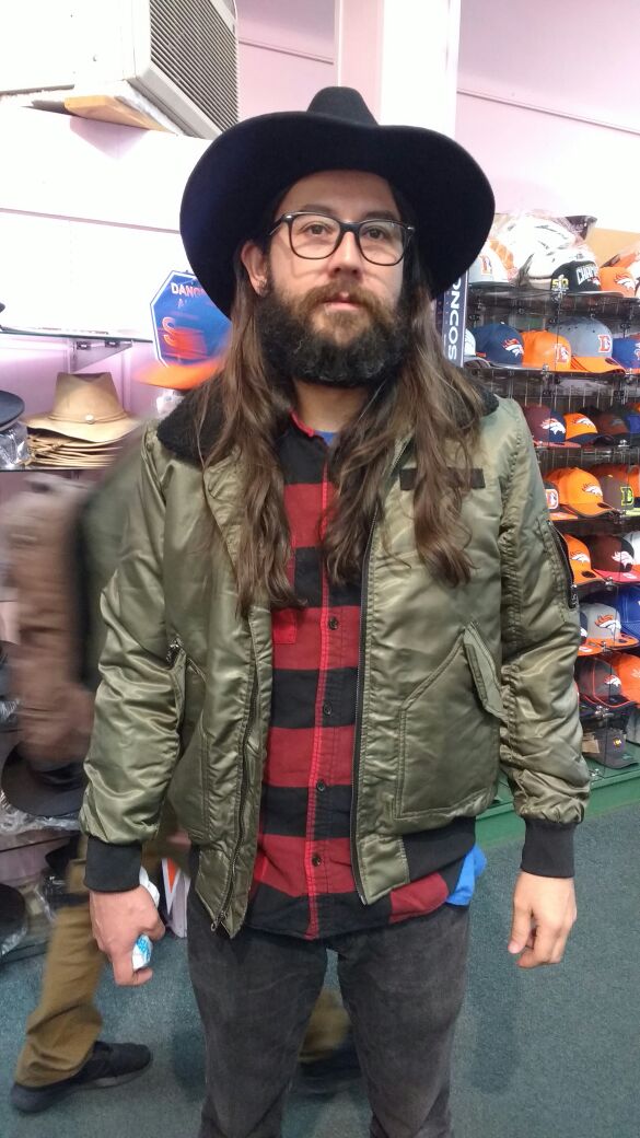 A bearded man with long hair poses for the camera wearing a black cowboy hat, black rimmed glasses, and a red and black flannel shirt. 