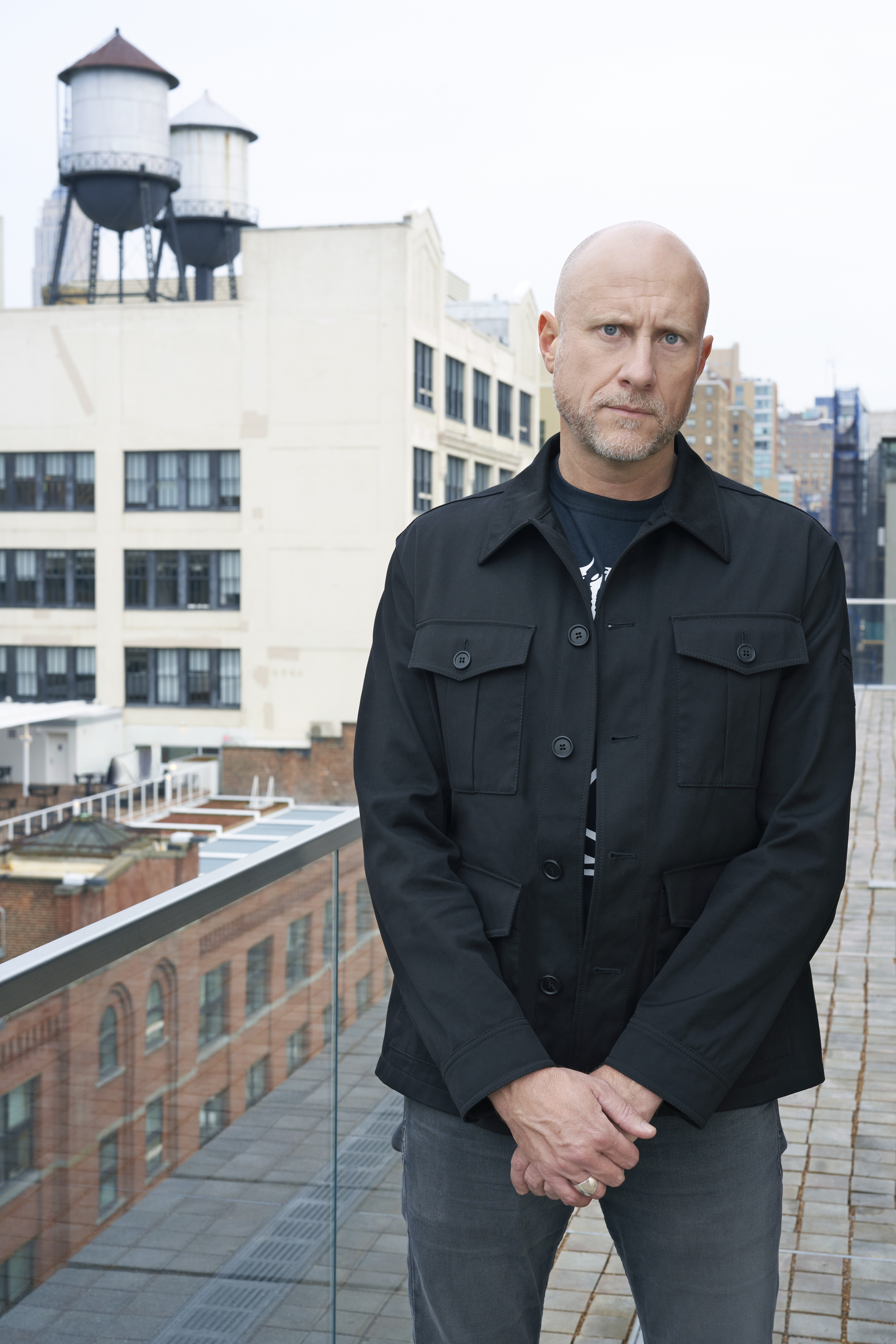 Portrait of Trevor Paglin standing against an urban landscape. He is wearing a dark coat as his cobalt eyes look sternly into the camera. .