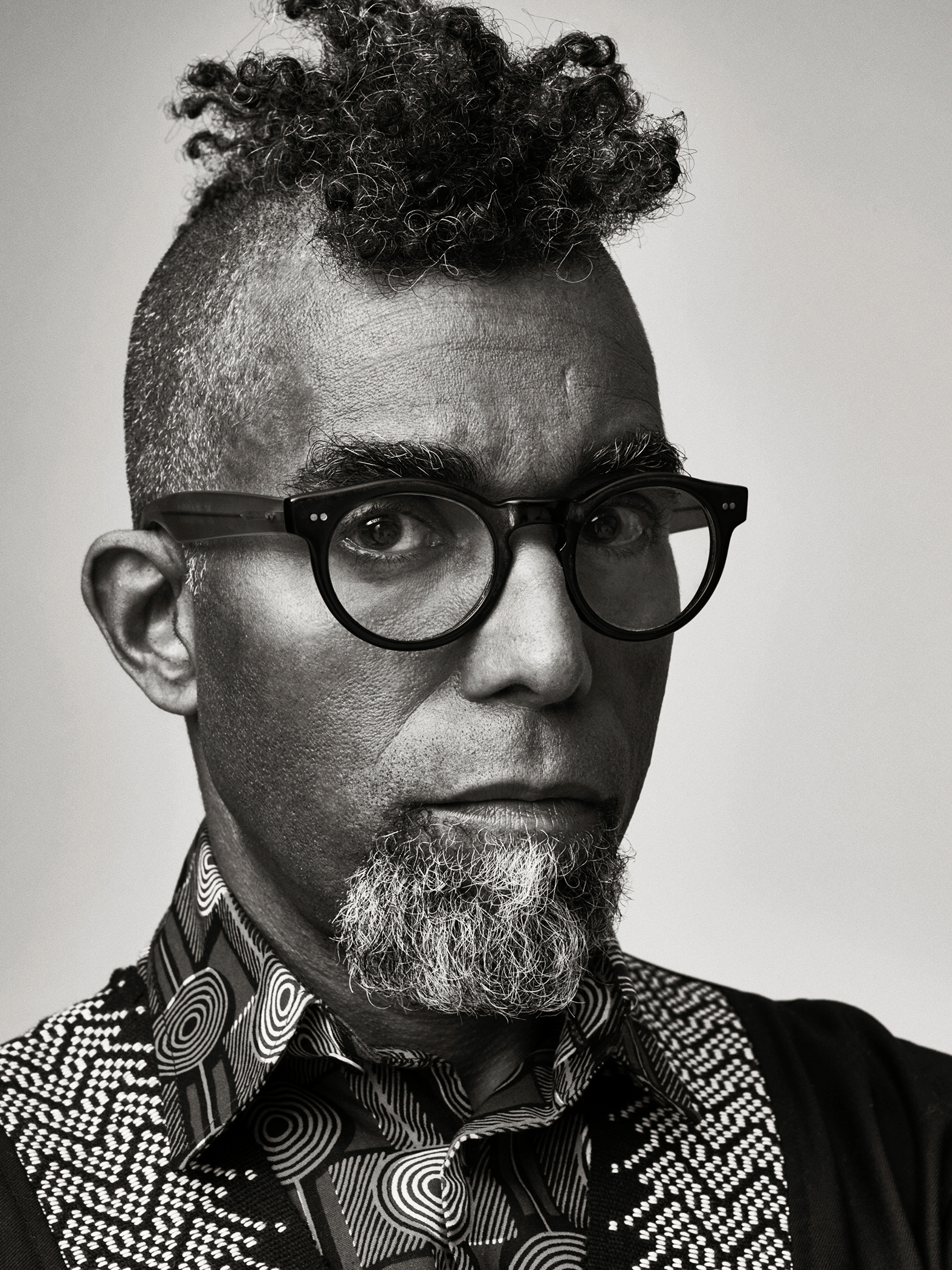 A black and white portrait of Dread Scott from the shoulders up against a neutral gray background. He looks seriously into the camera through his black rimmed glasses as a circular-patterned collar frames his salt-and-peppered beard and a tight black curly mohawk defines his upper face.  