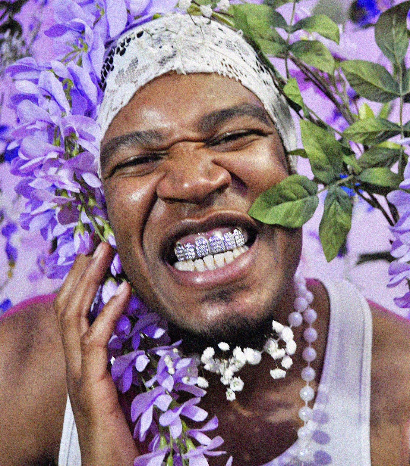A portrait of Tyriq, wearing a white tank top and what looks like a lace headband, surrounded by purple flowers and greenery. All of Tyriq’s teeth are showing, and it appears as though there is purple jewels on his teeth. 