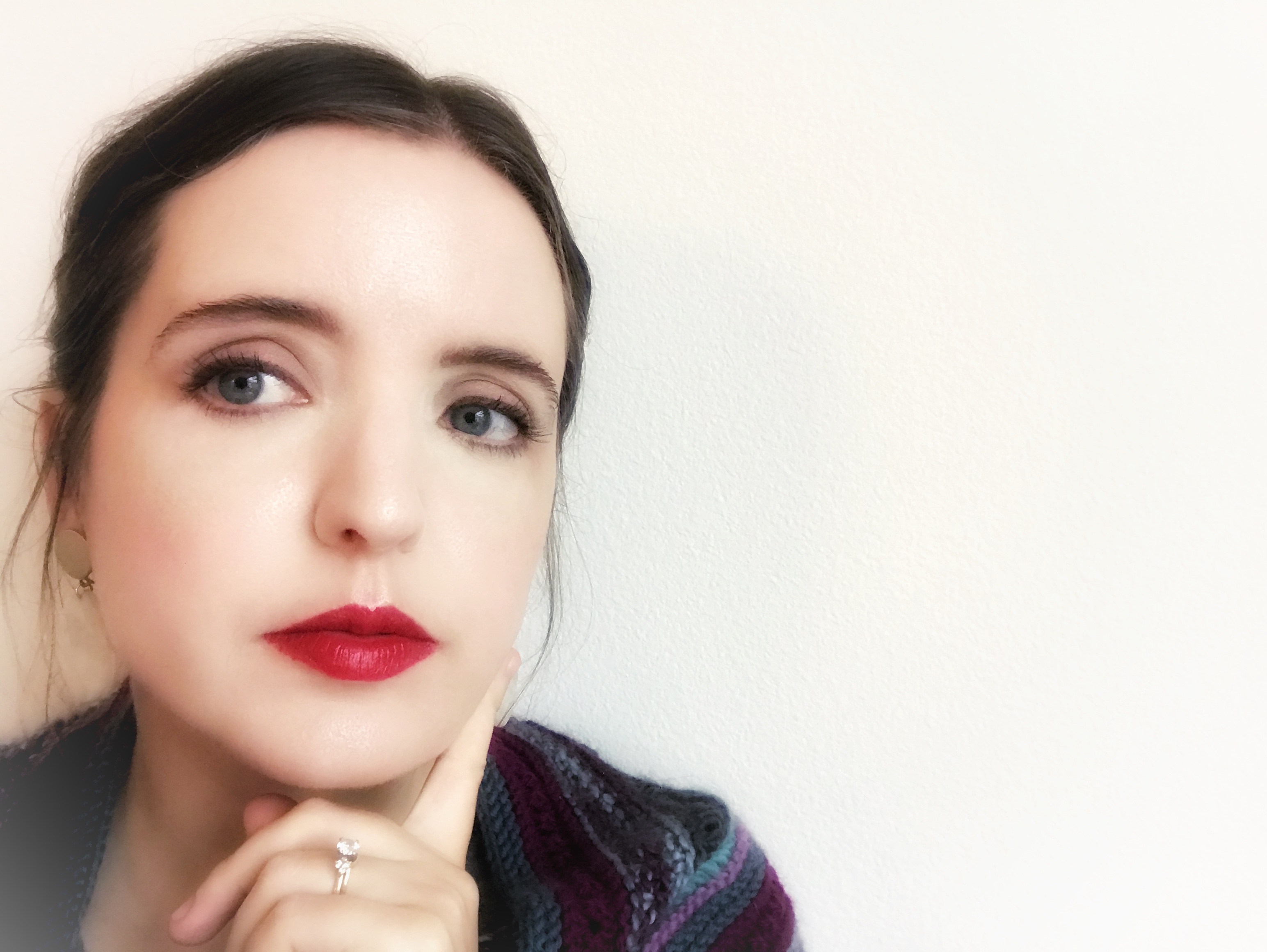 Close up selfie of visual artist, writer, and educator Erin Schalk in front of a white textured wall. Her skin is glowing, her eyes are focused elsewhere, and she has bright red lipstick and black eyeliner on. Her brown hair is up in a loose style and one finger in gently up near her face. 