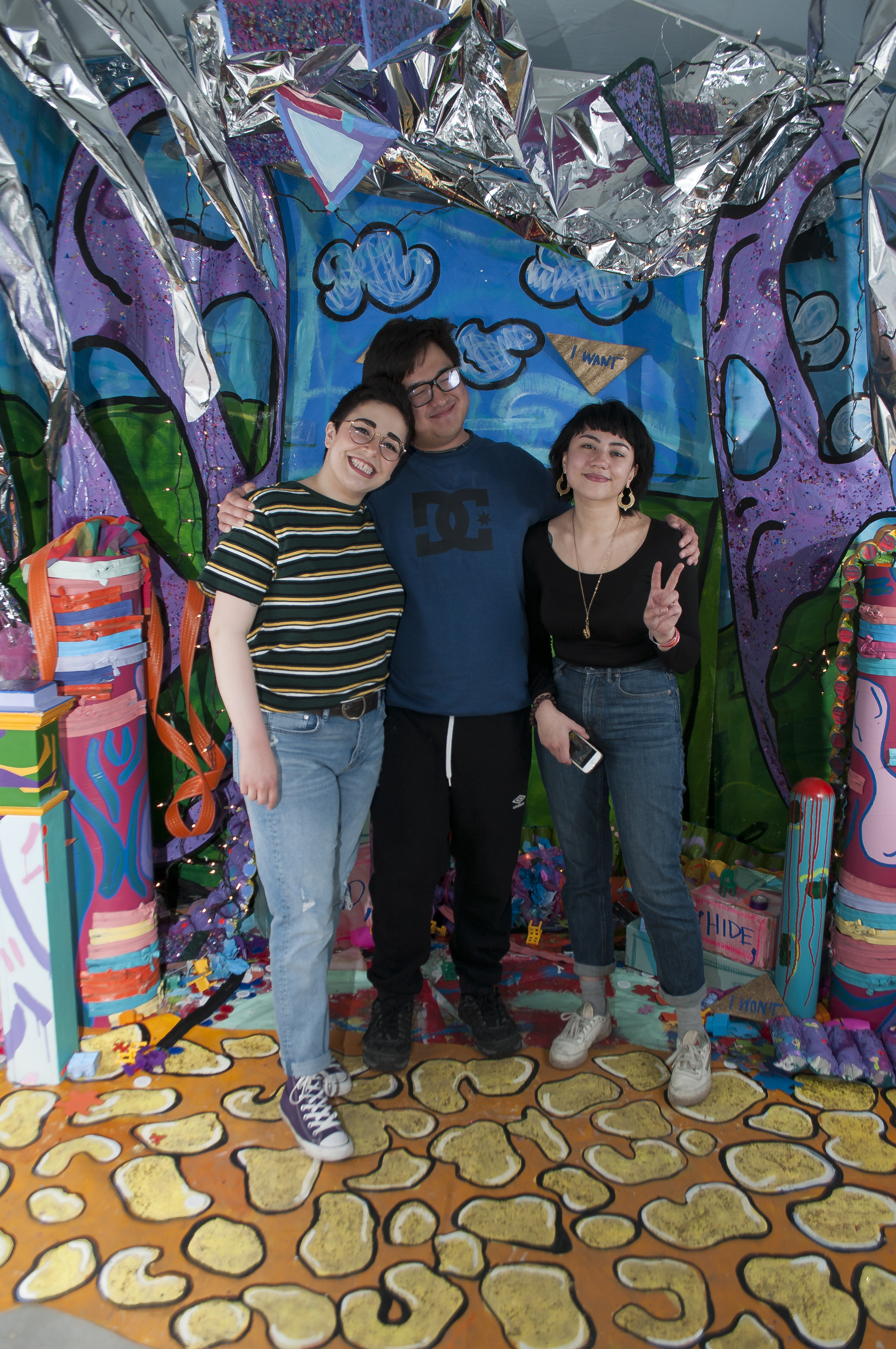 A group of three young adults smile warmly in front of a colorful set. 