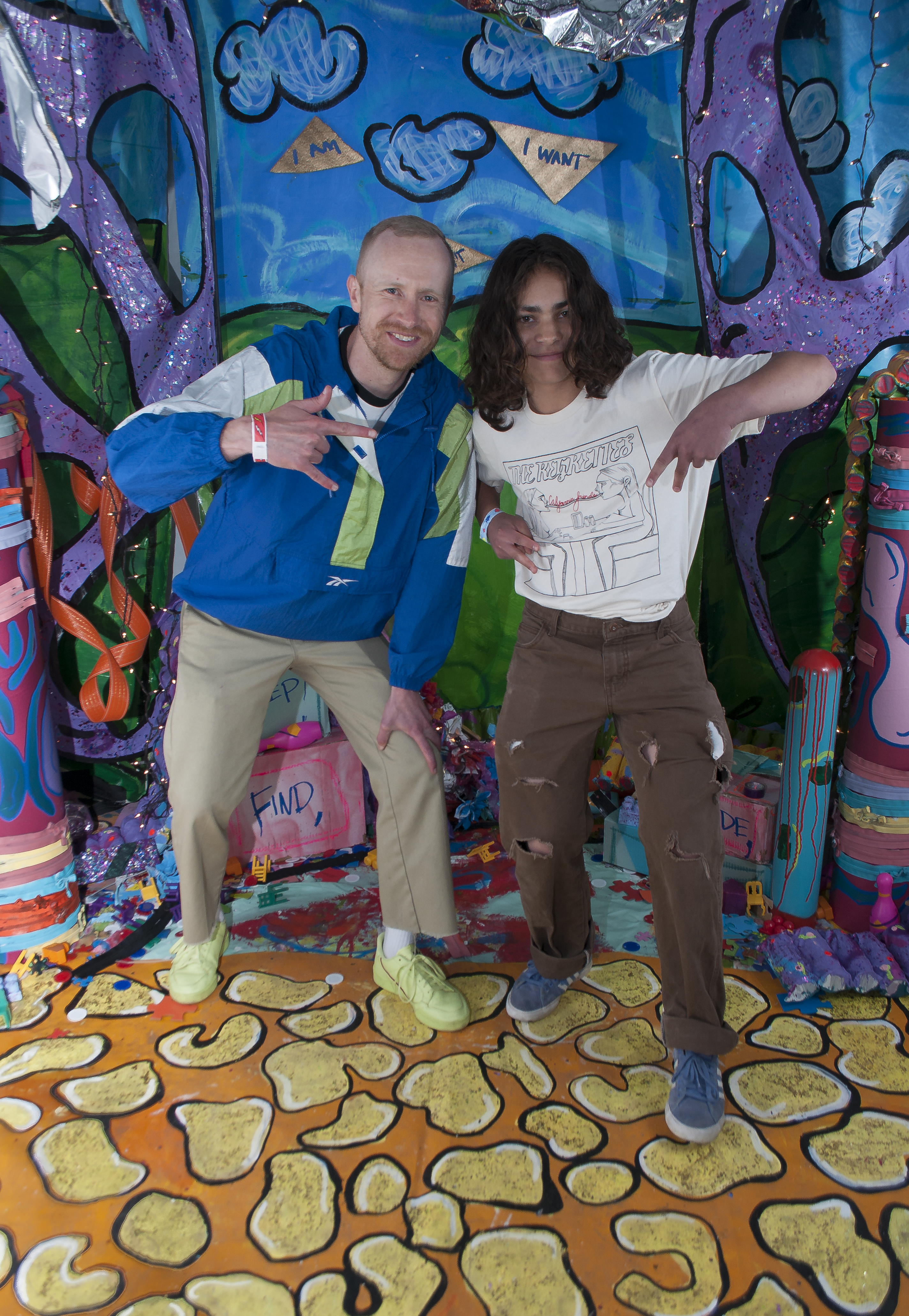 Two young adults pose in front of a colorful set. They are flashing the peace sign while smiling warmly into the camera. 