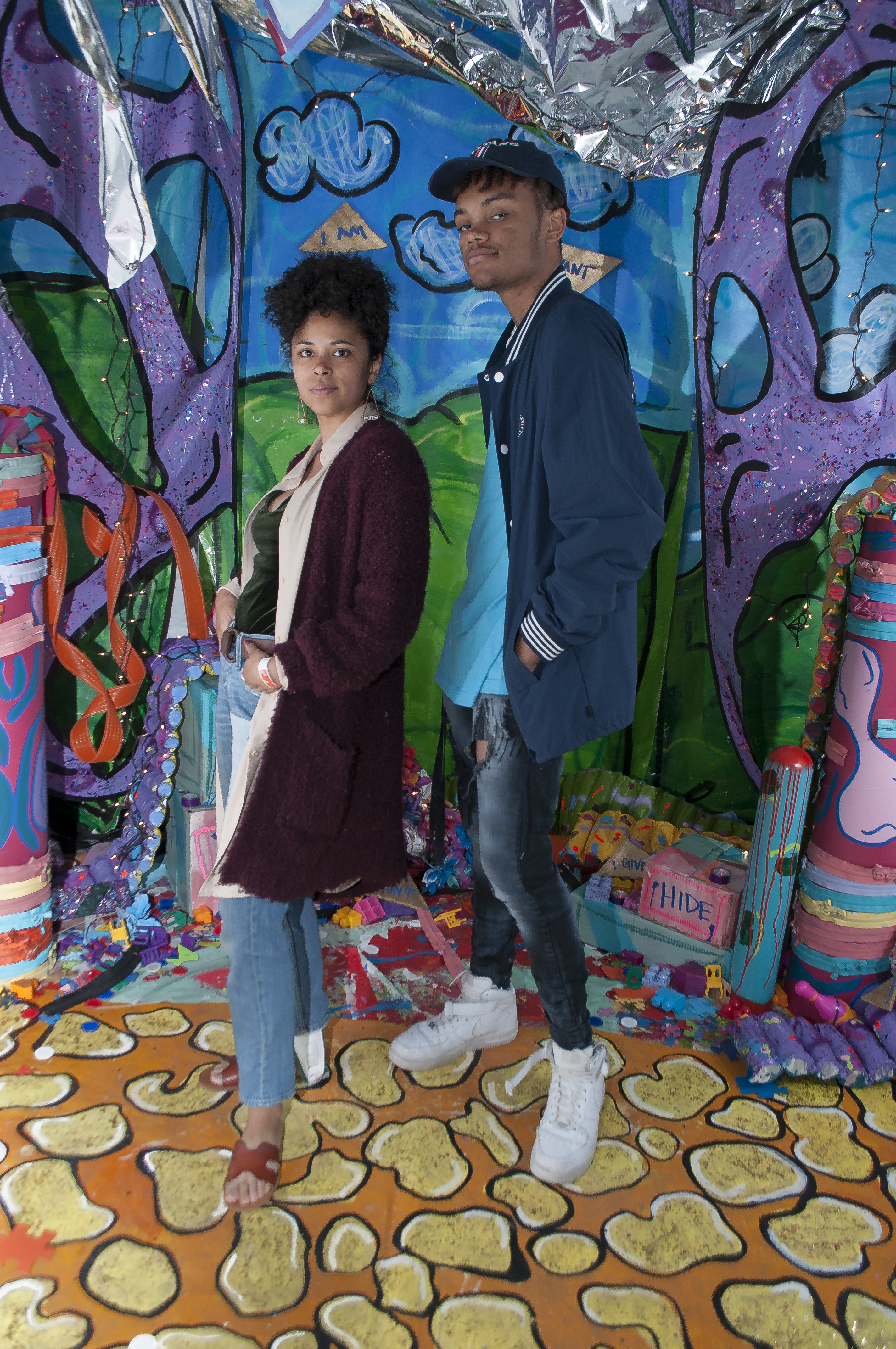 A young man and young woman pose in front of a colorful set. They pose confidently for the camera with neutral expressions. 