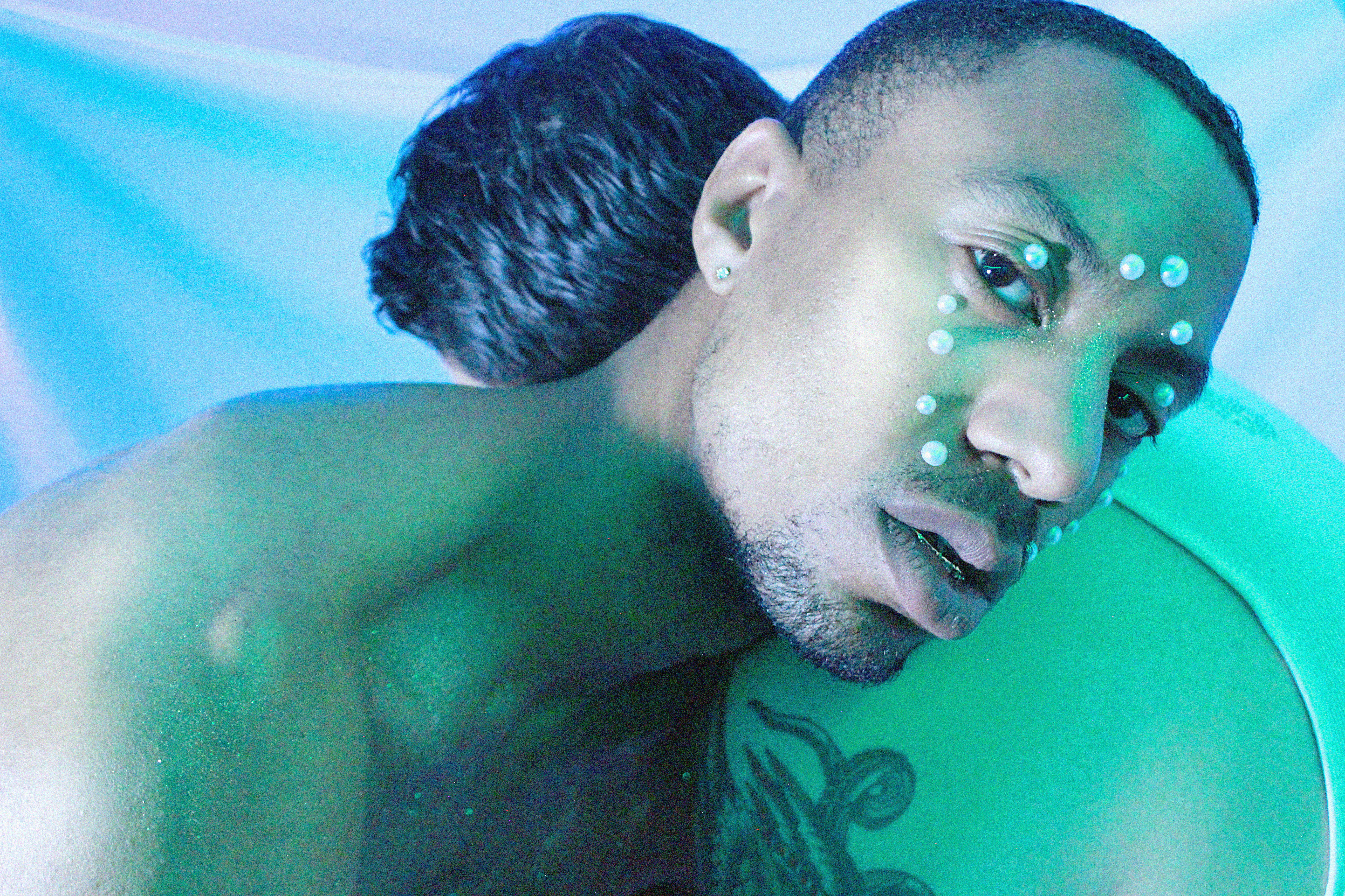 A photo of two men facing one and laying on one another’s shoulder. Only one of their faces is in view, and it’s covered in pearls. The photo looks as if it was taken underneath a blue/green hue and the backdrop is comprised of the colors blue, purple, and green.