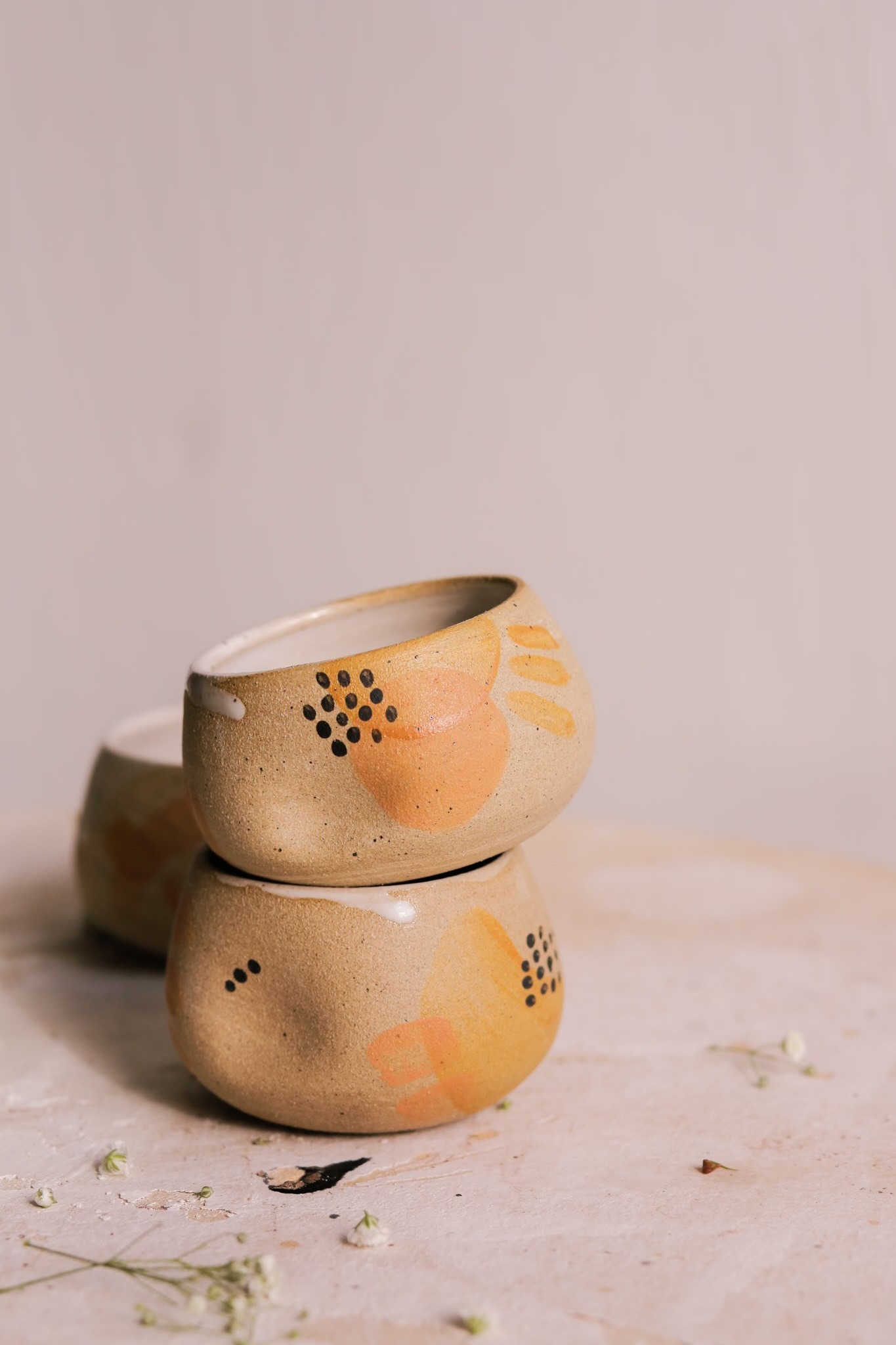Two small mugs stacked on top of one another, a third one is situated behind the two. The mugs are on a textured surface and in front of a dark cream colored backdrop. The mugs are neutral colors with black and orange abstract shapes on them. 