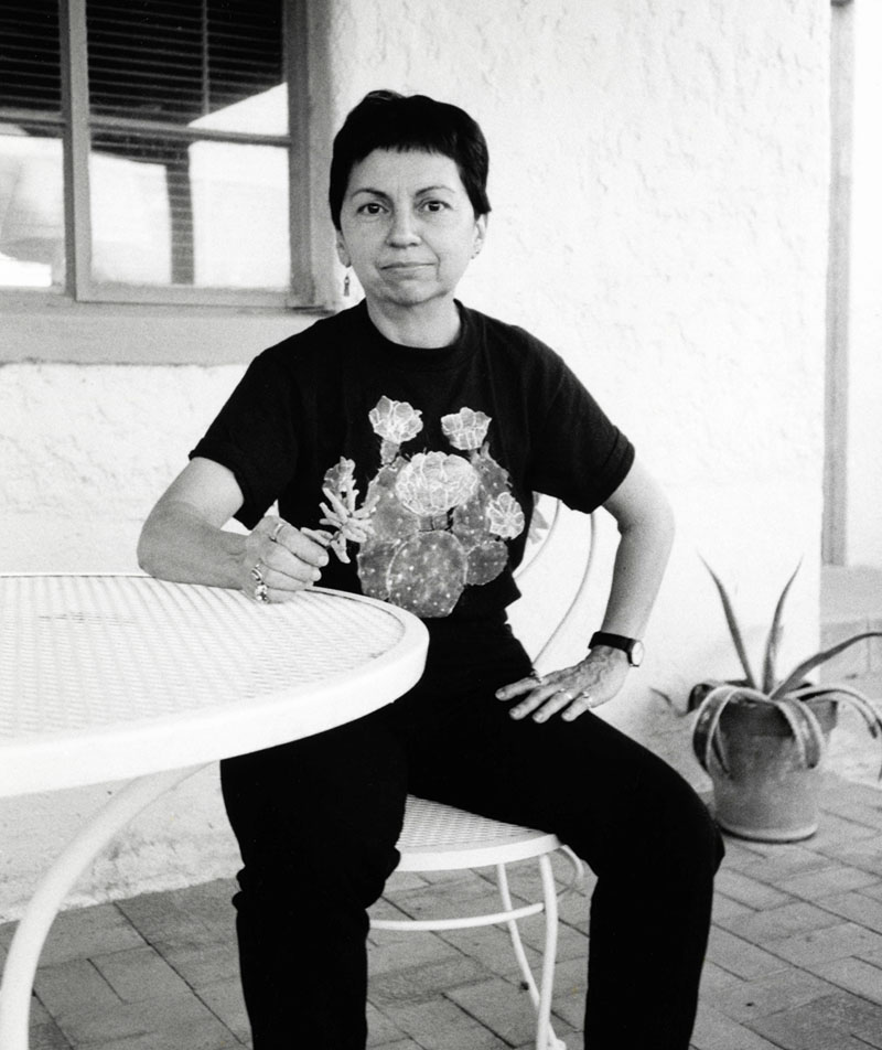 black and white photo of Gloria Anzaldua sitting outside at a table, fist on table.