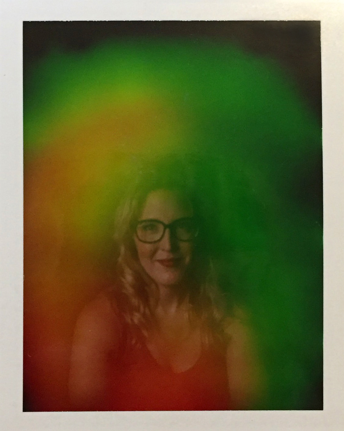 A polaroid photo of Heather, surrounded by a green, orange and red aura. She is wearing glasses and smiling softly. 