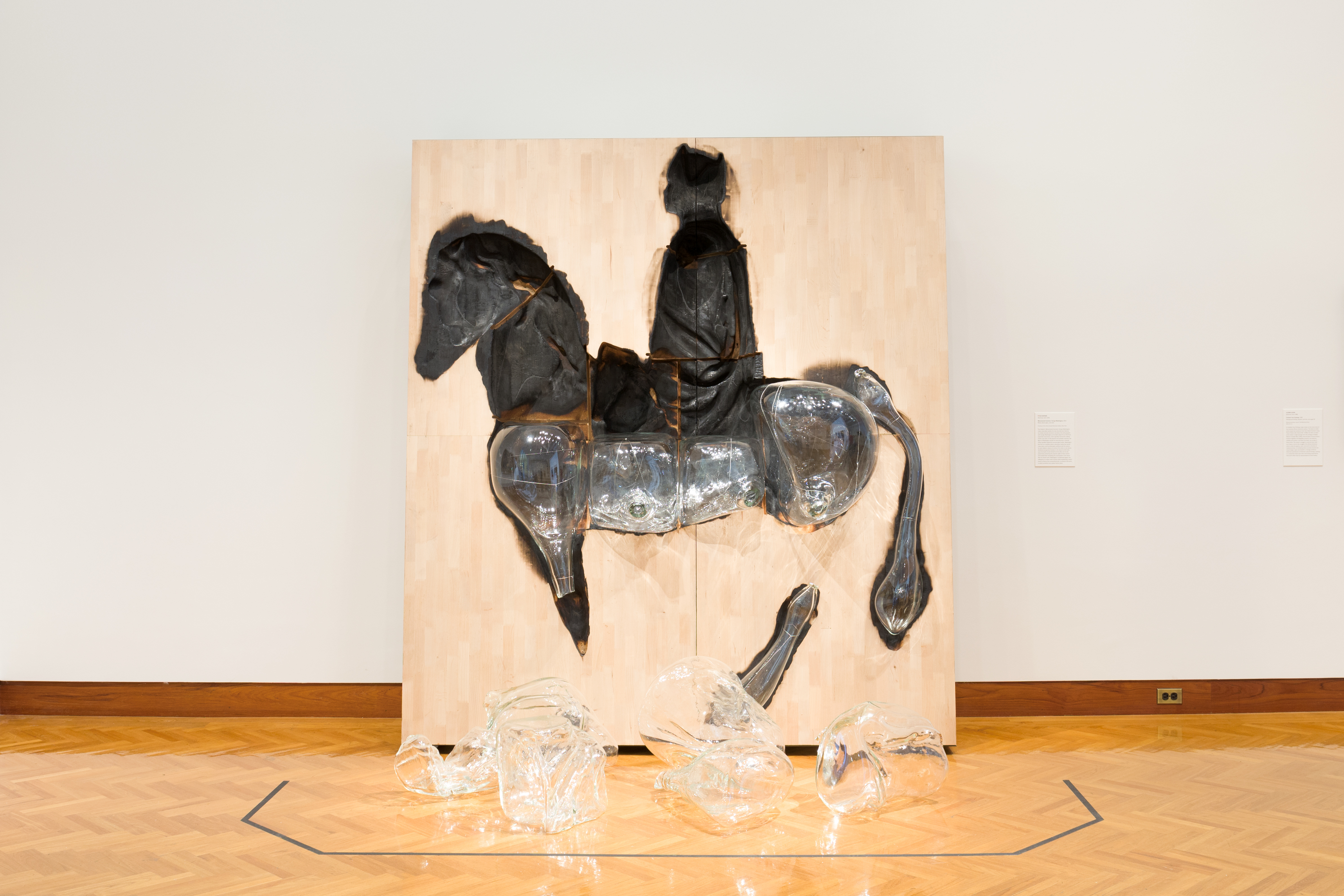 Titus: A large flat piece of wood rests against a wall in a gallery space. A figure of a man riding a horse is charred into the wood. Glass rests in the lower half of the charred spaces while on the floor of the gallery remains the upper portion of the horse and man.