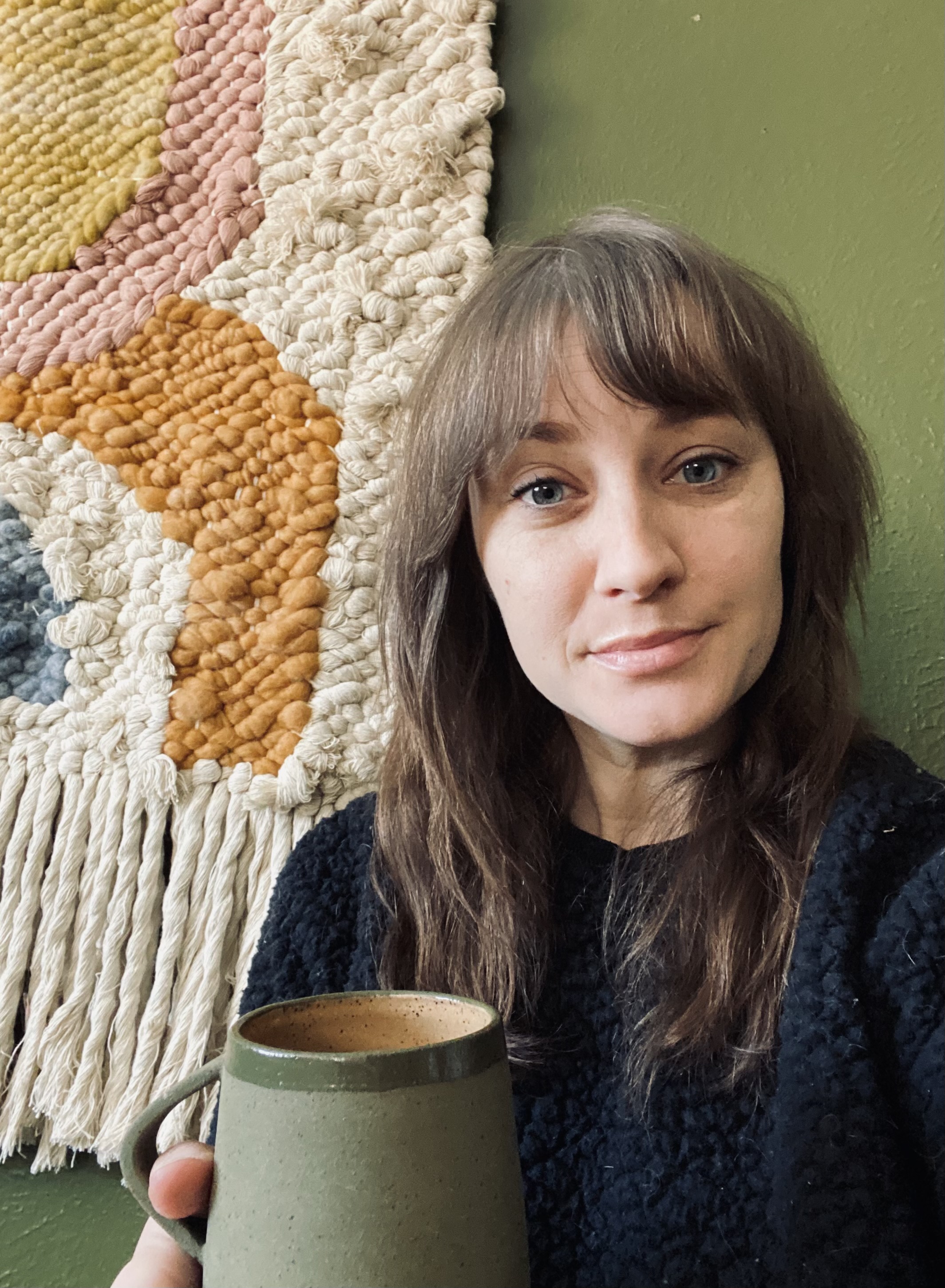 selfie of artist Natalie Legg smiling softly with her mouth closed and sporting medium length brown hair with fringe. She’s wearing a black sweater and is holding a forest green coffee mug. She’s standing in front of a large multi-color woven wall hanging that is hung on a green wall. 