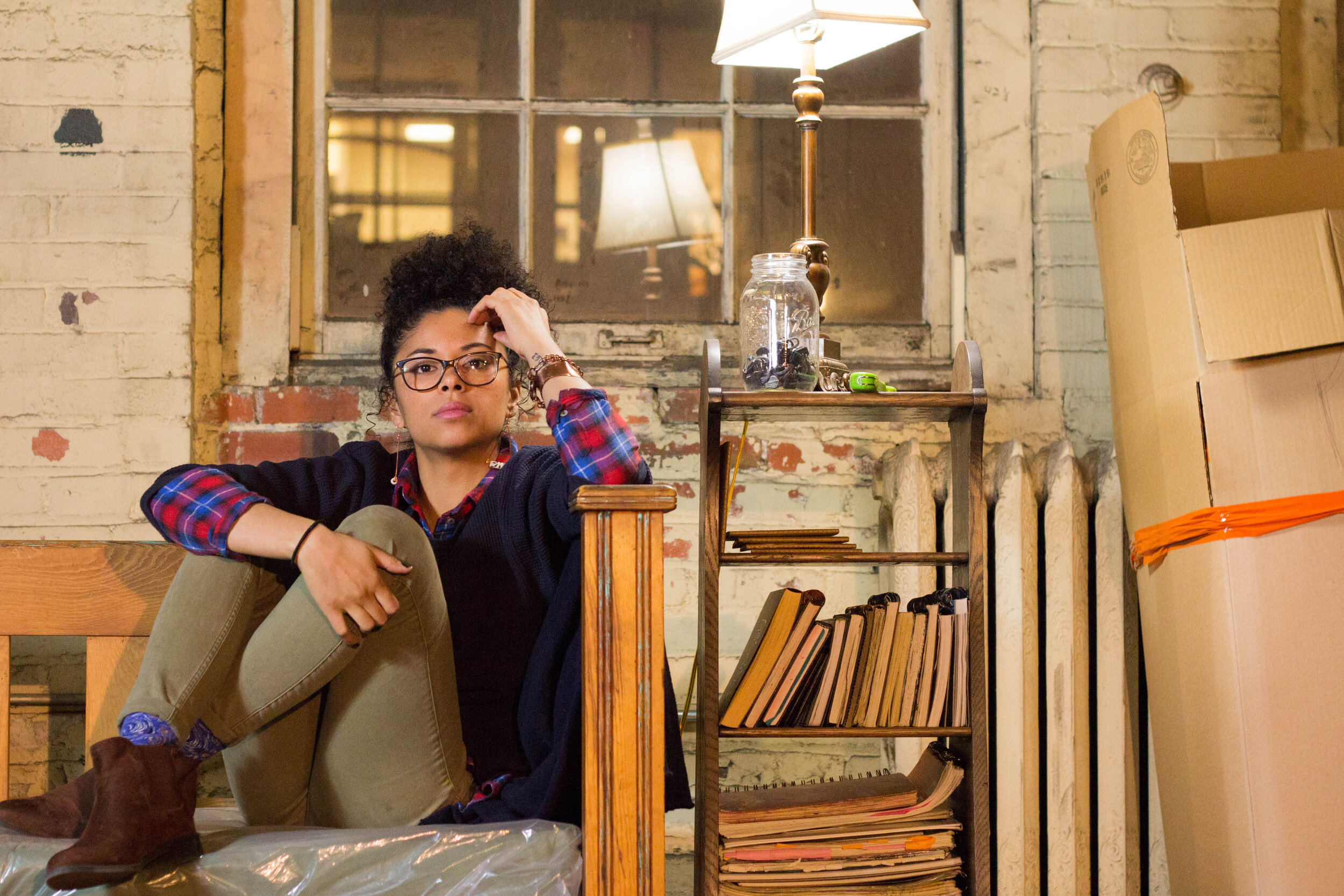 A portrait of Moe Gram sitting in an industrial building next to stacks of books and a table lamp