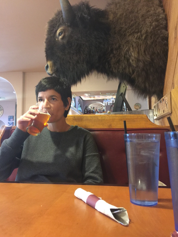 Cynthia sits at a restaurant table drinking a light beer. There is a mounted bison head on the wall behind her. 