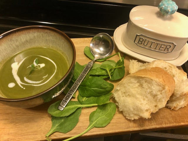 A bowl of green soup with white sauce is on a cutting board surrounded by spinach leaves. 