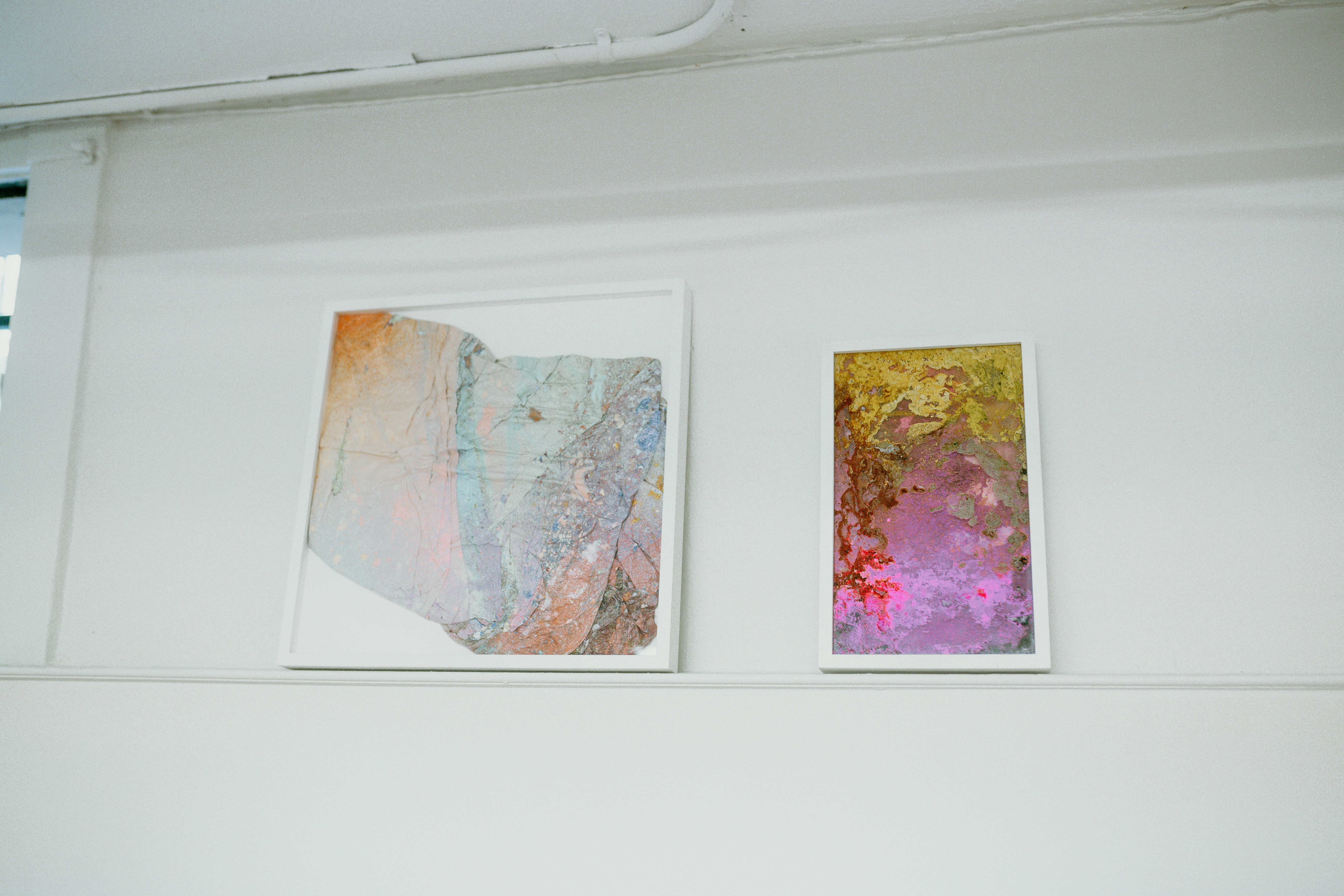 Two abstract artworks on a white wall. They are medium sized and have pinks, yellows, and beiges. 