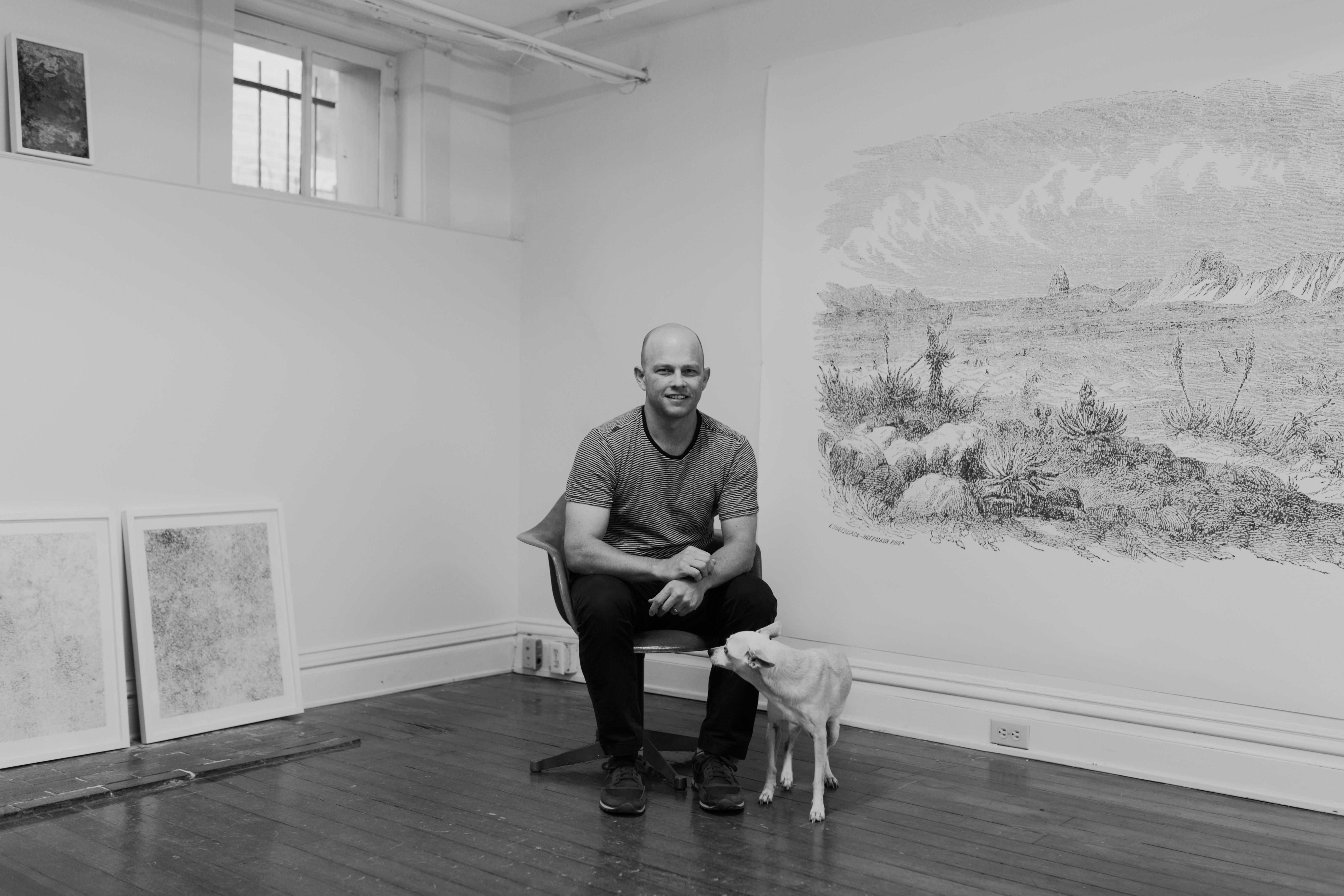 Portrait of Chris Oatey sitting in an indoor space. There is a medium sized white dog by his side. On the wall next to him is a large artwork of an arid landscape. ﻿