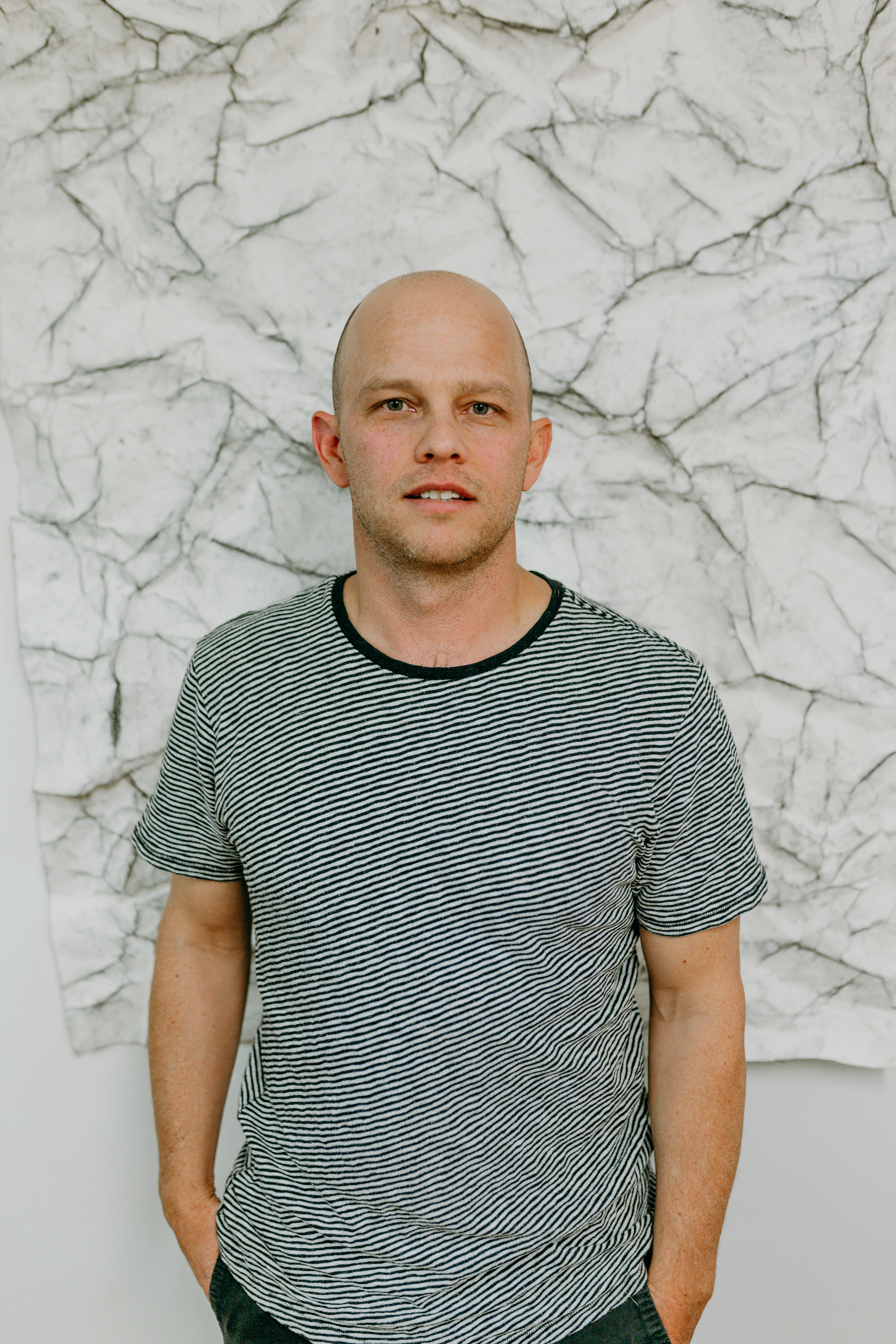 Portrait of Chris Oatey in front of an abstract background resembling white cracked earth. He is bald, wears a black and white striped t-shirt. His hands are in his pockets and he looks neutrally into the camera with his mouth slightly open. 