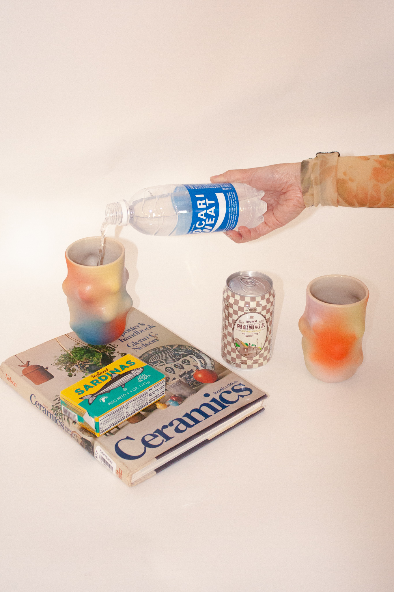 Objects photographed on a light pinkish, beige background. In view is a book about ceramics, and on top of the book is a lumpy, colorful mug. There’s a disembodied hand pouring water into the mug. Next to the book is a can and another lumpy, colorful mug.