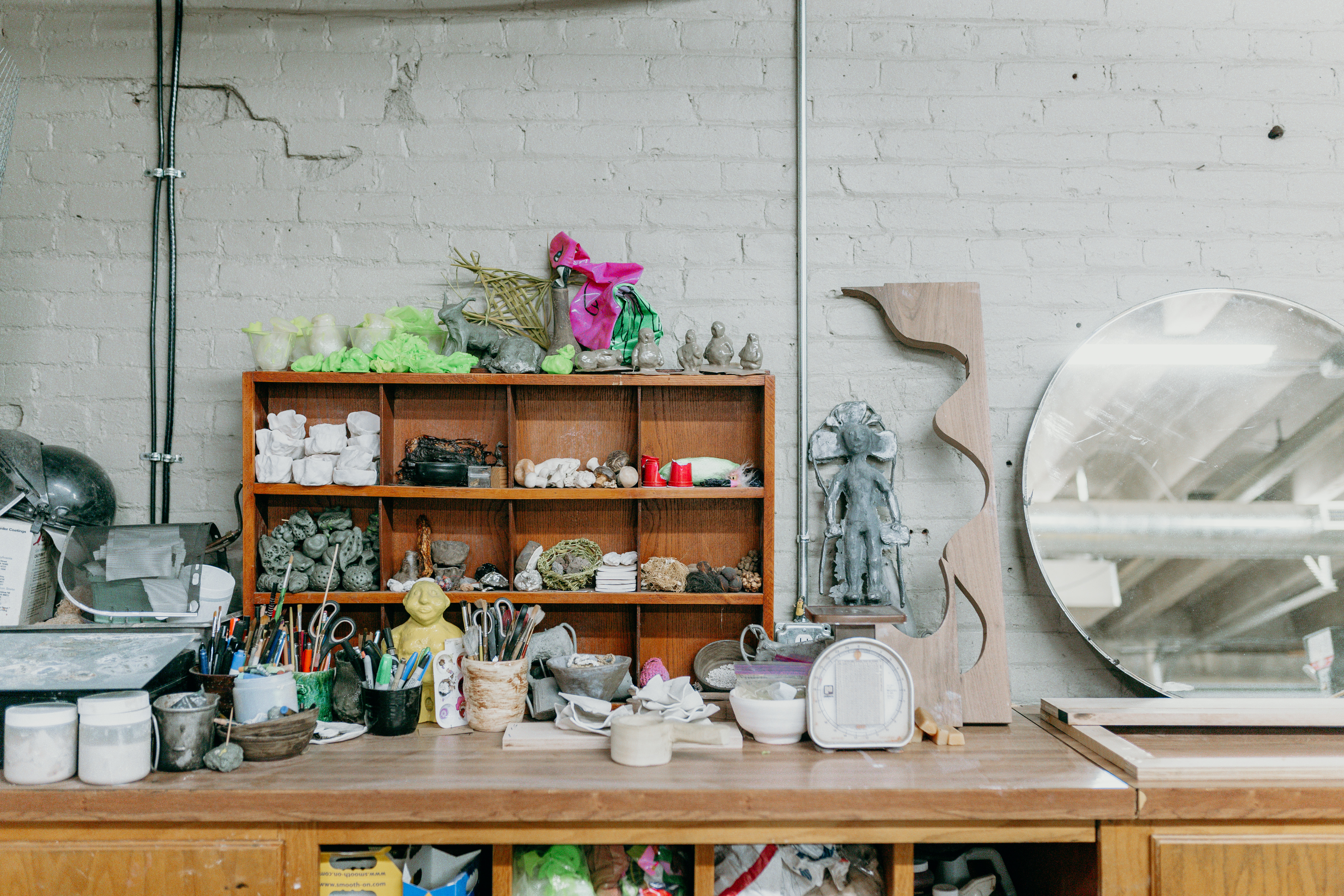 A workspace with several clay sculpting tools and work in progress sculptures. There is a mirror resting against a white painted brick wall. 