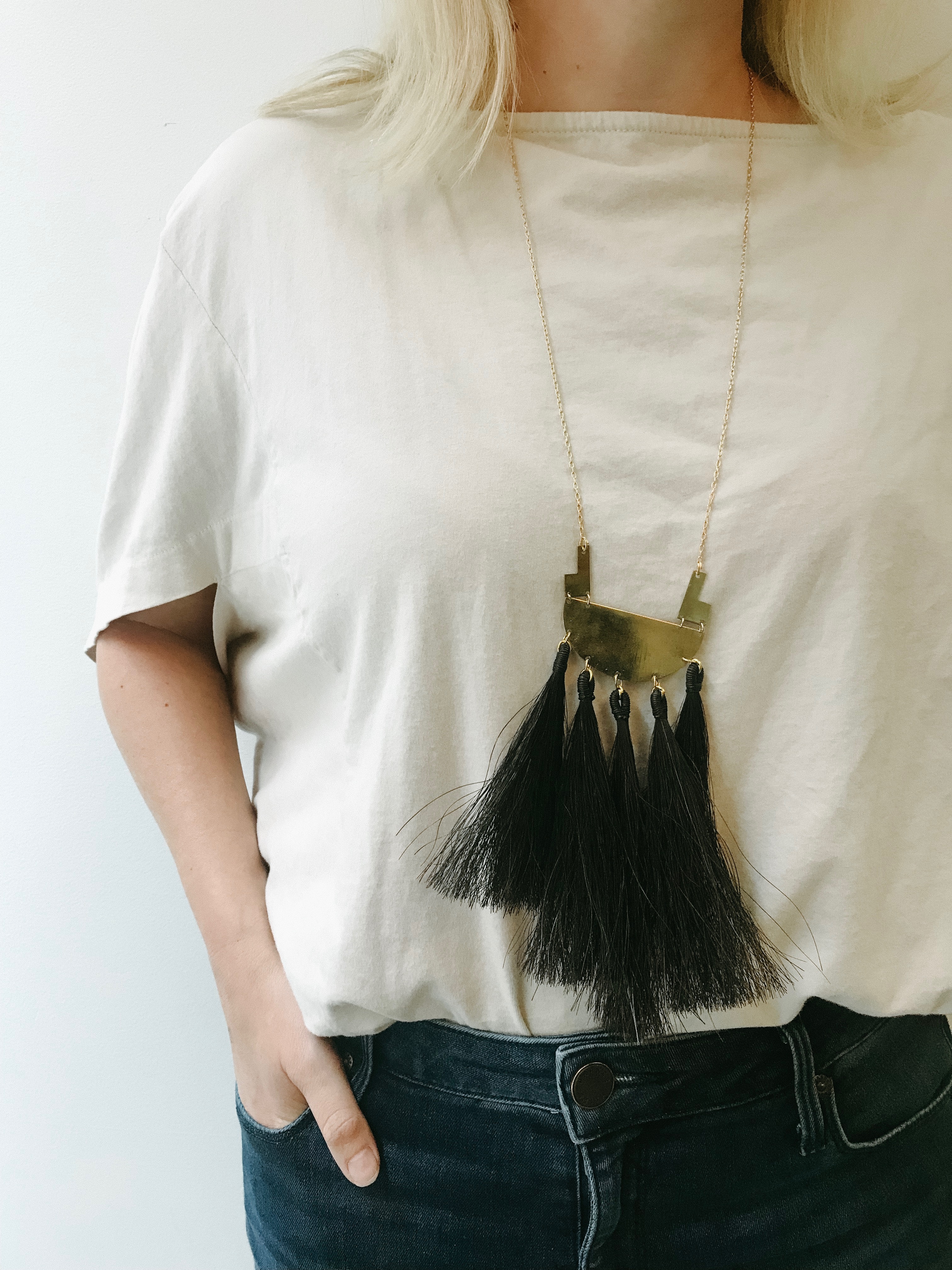 A woman models a large brass necklace. It hangs down to her mid torso and has black fringe. 