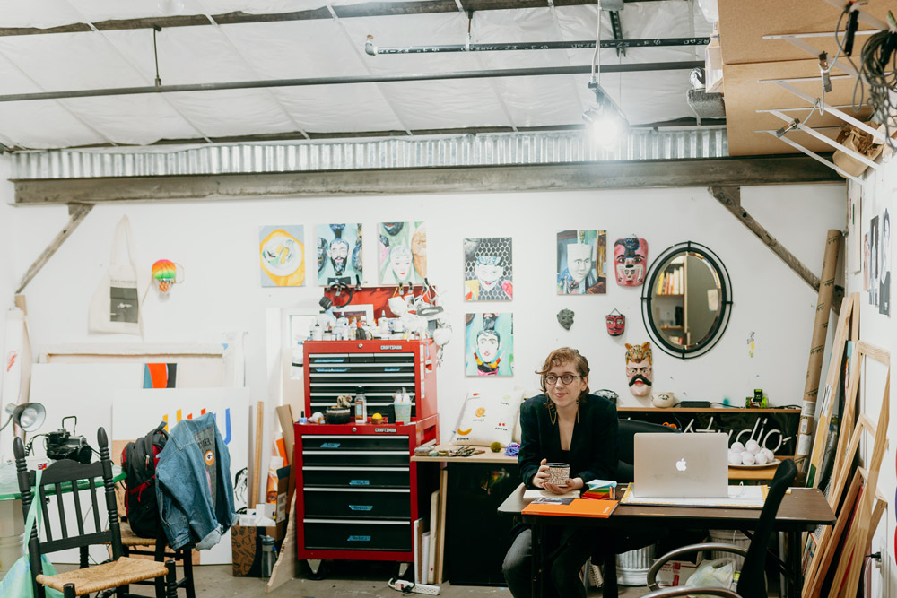 Sierra at her desk in her studio space. She is surrounded by several artworks. A mac book is open on her desk and she holds a ceramic mug in her hands. 