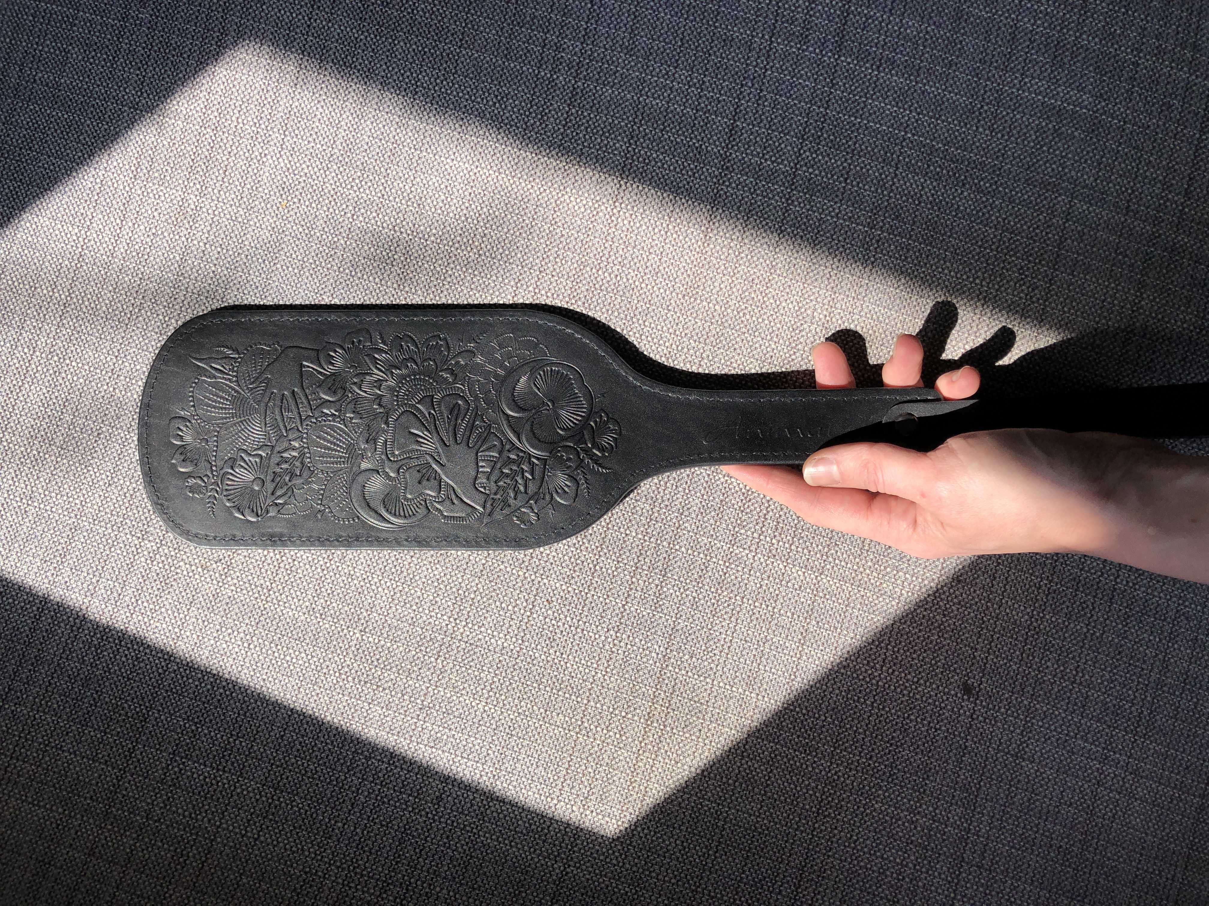A black leather paddle with an intricate design on it that features hands, flowers, and fruits. A hand is holding the paddle on a textured gray background. Light is seeping into the frame and creates a diamond shape, which frames the paddle. 