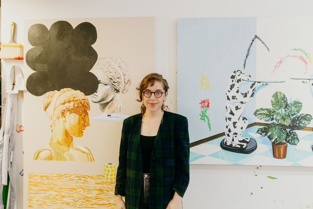 Sierra stands in front of two paintings. One depicts a dog sculpture holdling a platter next to a monstera plant. The other depicts two busts in a classical greek style. 
