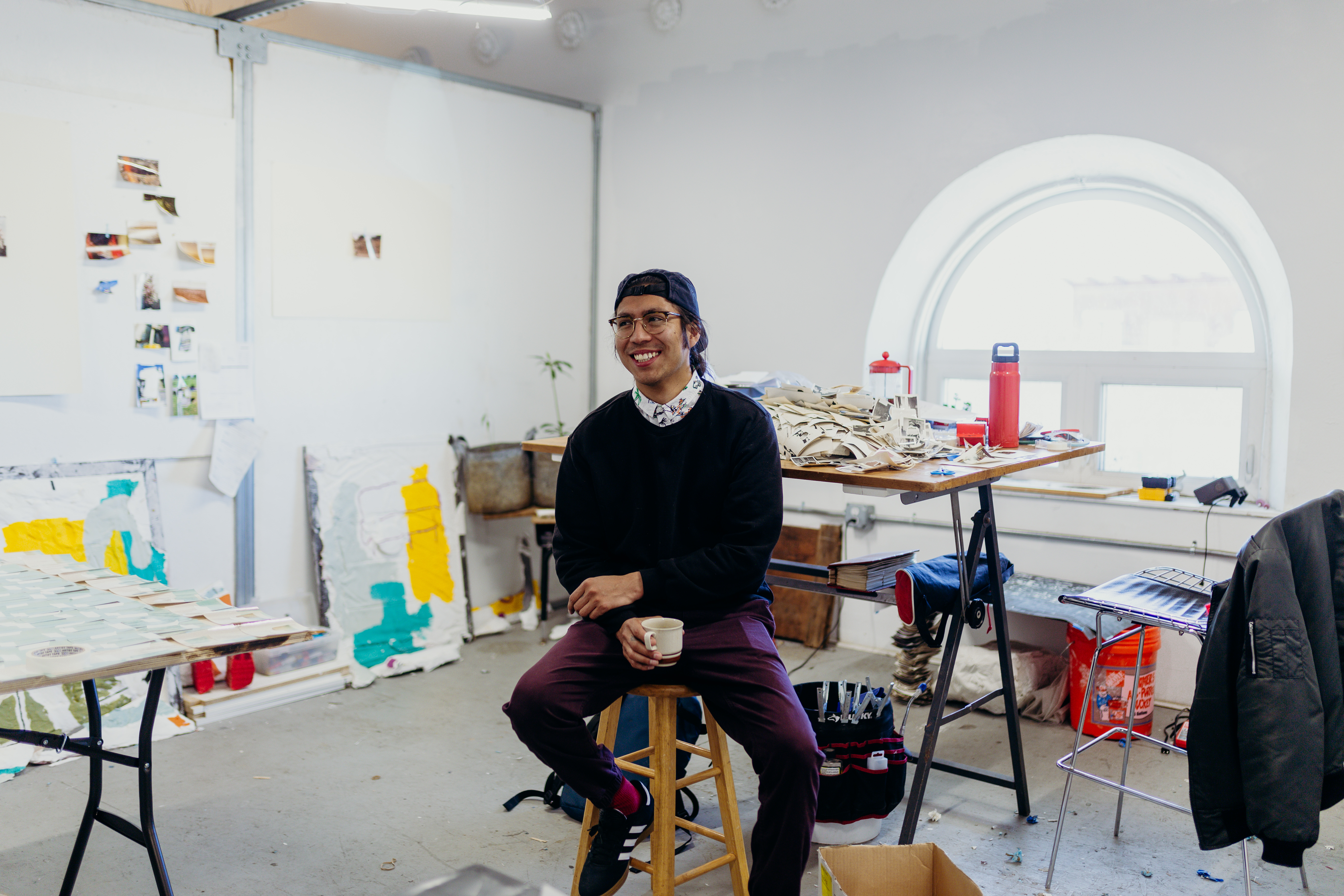 George P. Perez is sitting on a stool in his studio holding a mug and smiling at something off camera. Behind him is a table with various work materials. 