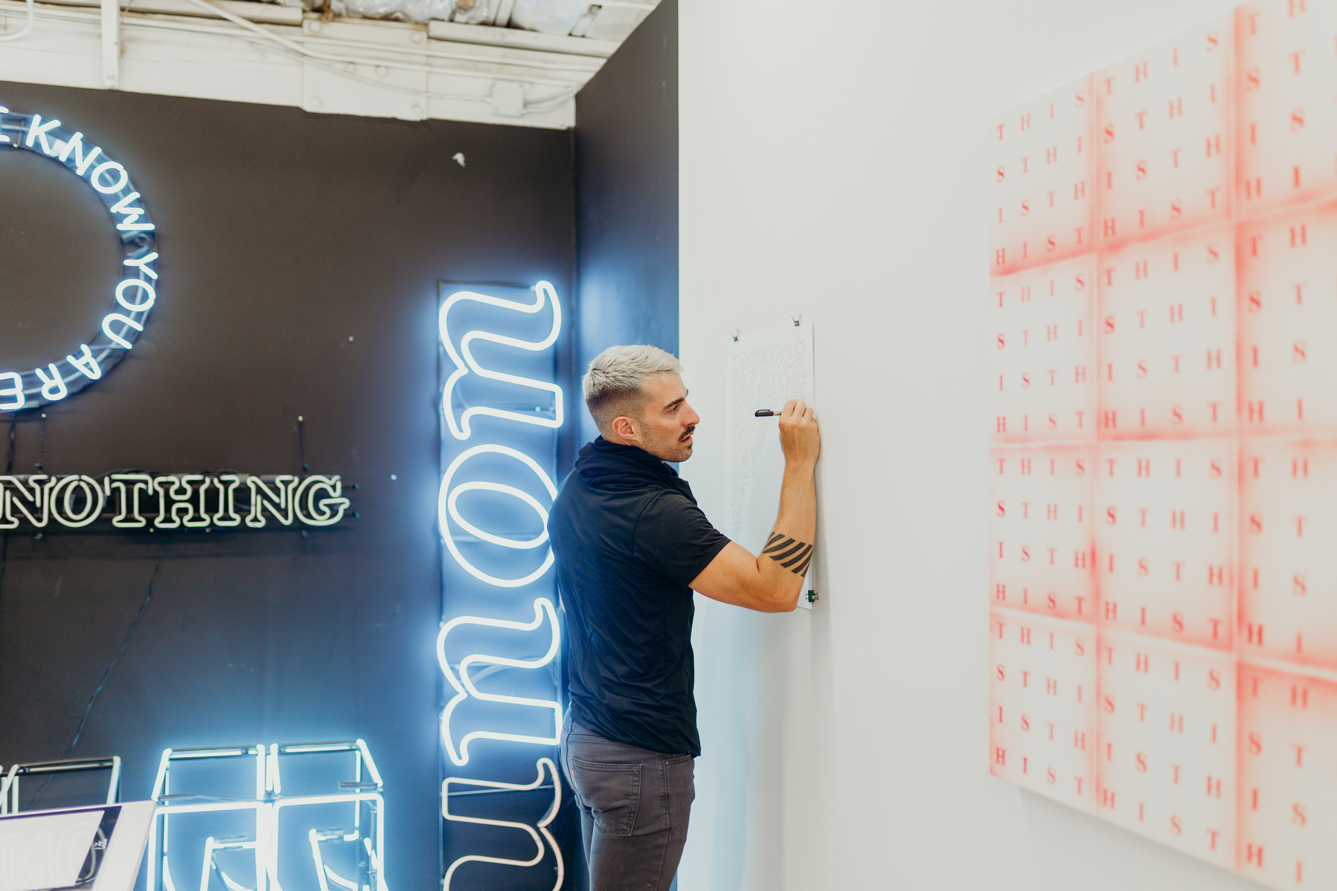 Wide candid shot of joel swanson drawing on a piece of paper hung on the wall. Behind him are several large neon artworks. . 
