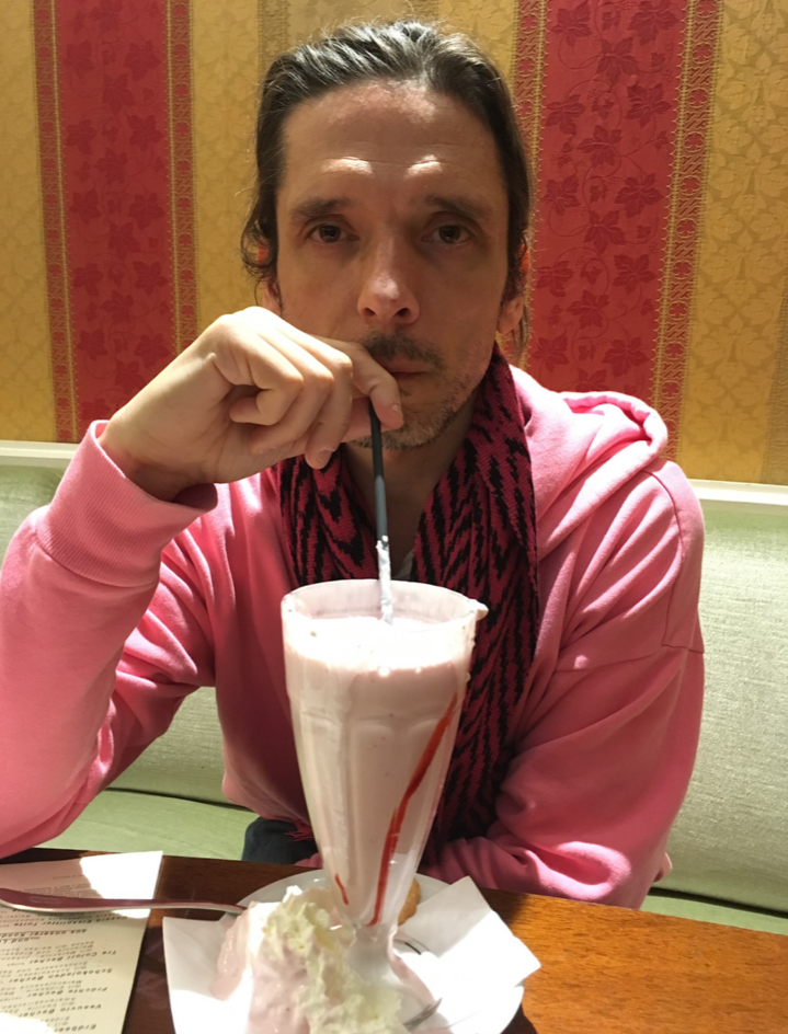  Portrait of Jeremy Deller leaning over a milkshake at a restaurant booth facing the camera against a red and yellow floral patterned wallpaper. His hair is pushed back and he carries a soft look on his face while wearing a pink hoodie with a pink and black zig-zag scarf.  He is holding the straw of the milkshake to his mouth with his right hand. 