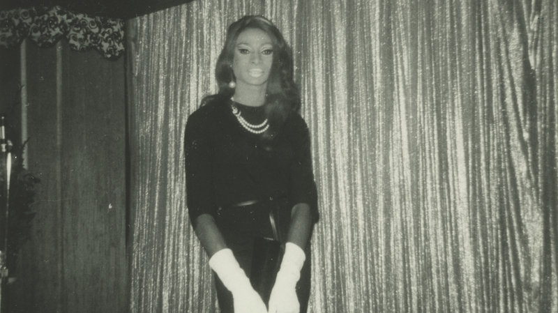 Vintage black and white photo of Jackie Shane in white elbow length gloves and black dress standing facing the camera in the center of the horizontal photo. 
