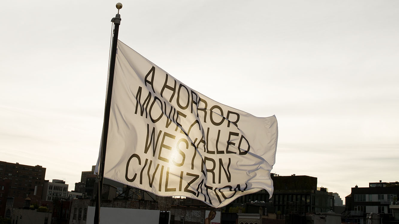 Jayson Musson, A Horror, 2017. A white flag with the text “A HORROR MOVIE CALLED WESTERN CIVILIZATION” in all capital letters flies against an urban landscape. The sun is shining through the material of the flag. 