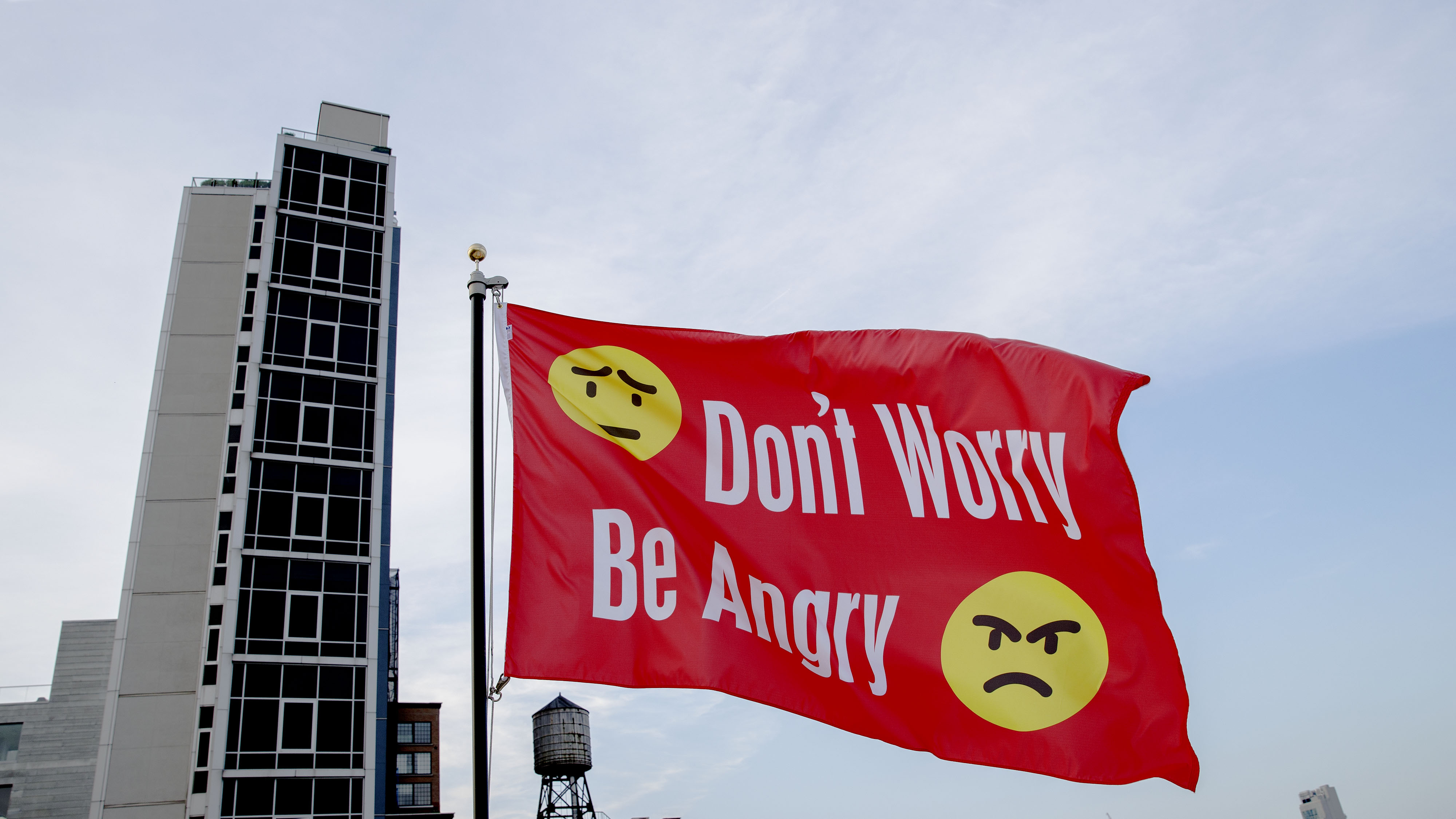 Jeremy Deller, Don’t Worry Be Angry, 2017. Red nylon flag reading "Don't Worry Be Angry" in white text. There are two yellow emoji faces; one is worried and the other is visibly angry. 