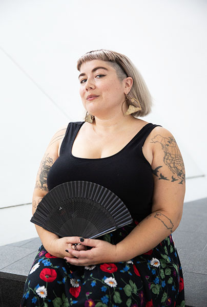 portrait headshot of Lares Feliciano. Sitting in a floral skirt, black tank top holding an open black hand fan.