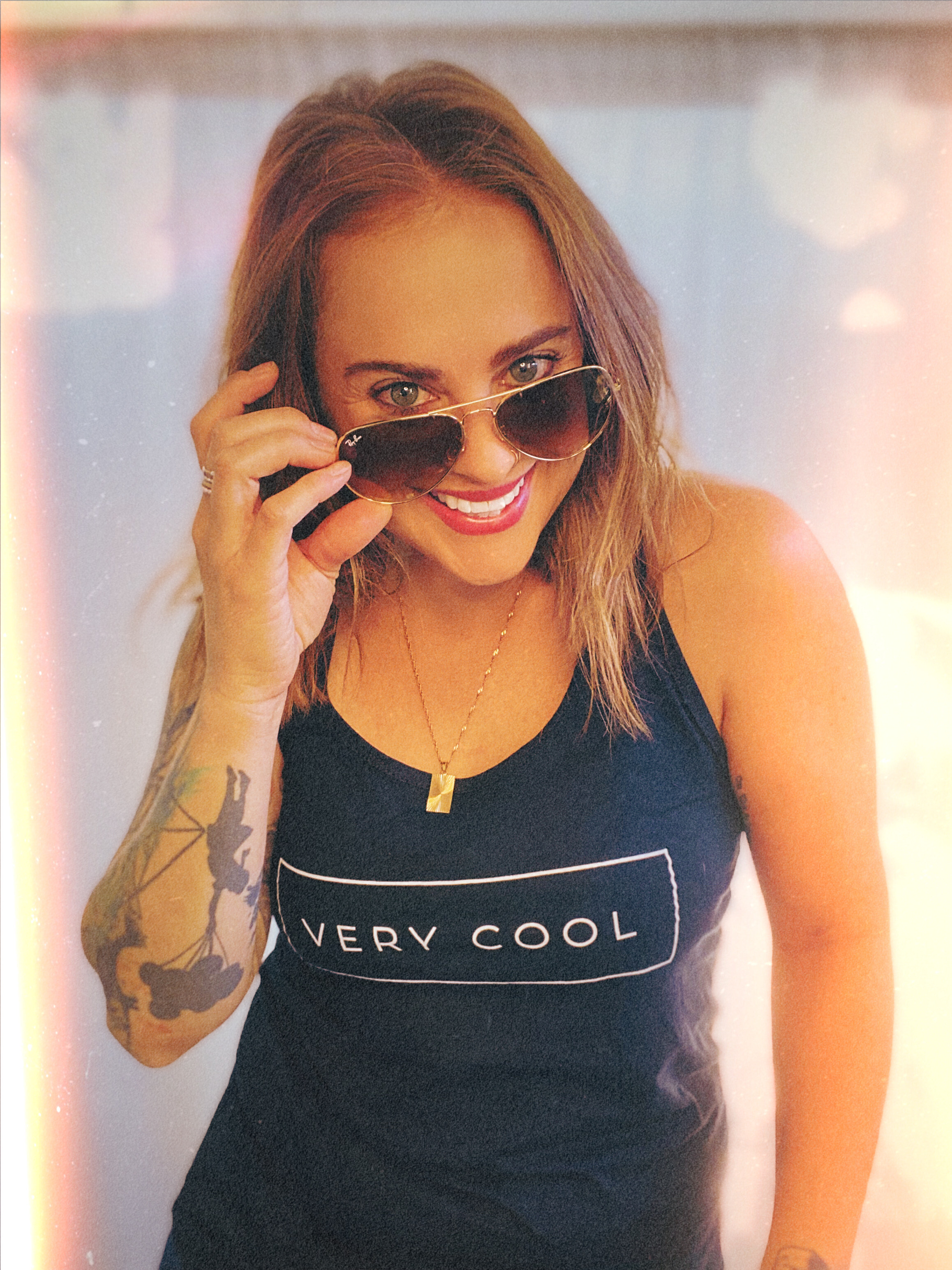 Portrait of Cassandra of Lux + Luca jewelry line, wearing a black tank top that says “very cool” and aviator sunglasses. Cassandra is tilting the sunglasses down and smiling at the camera. 