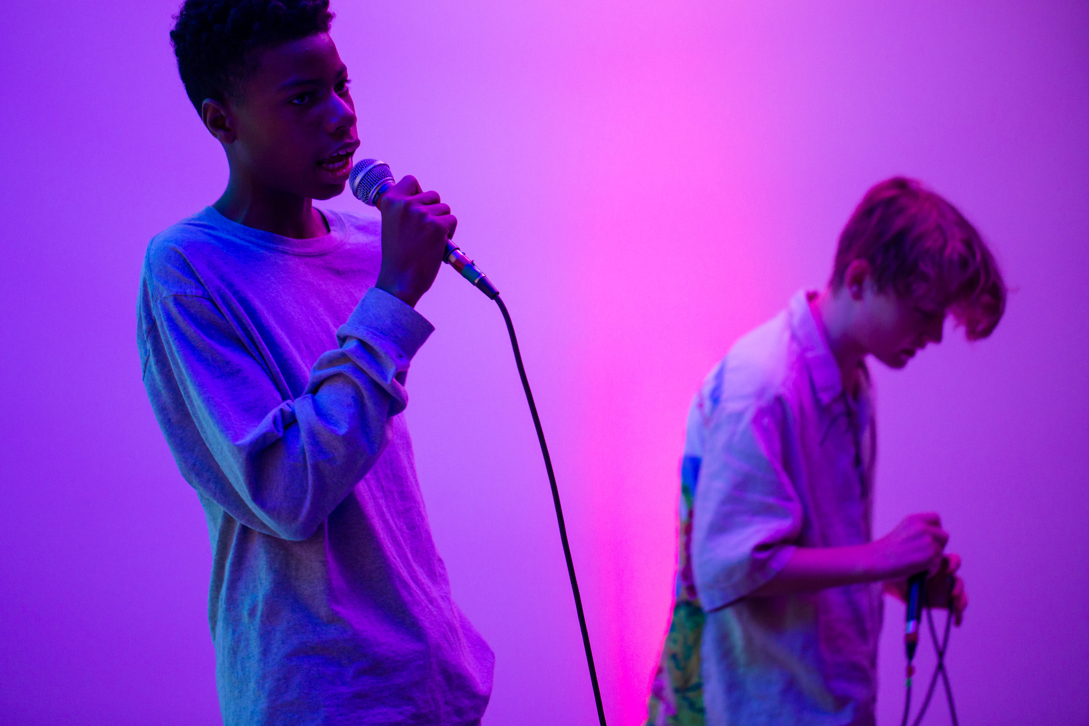 Two young men are performing and holding microphones. They are light in purple and pink. 
