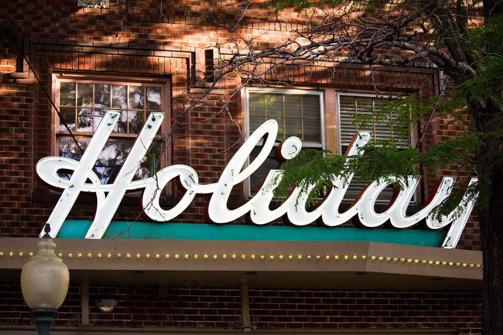 Close up street view of a sign on a brick building that reads “Holiday” in white, cursive lettering.  Behind the sign are three windows, and below are marquee lights. 