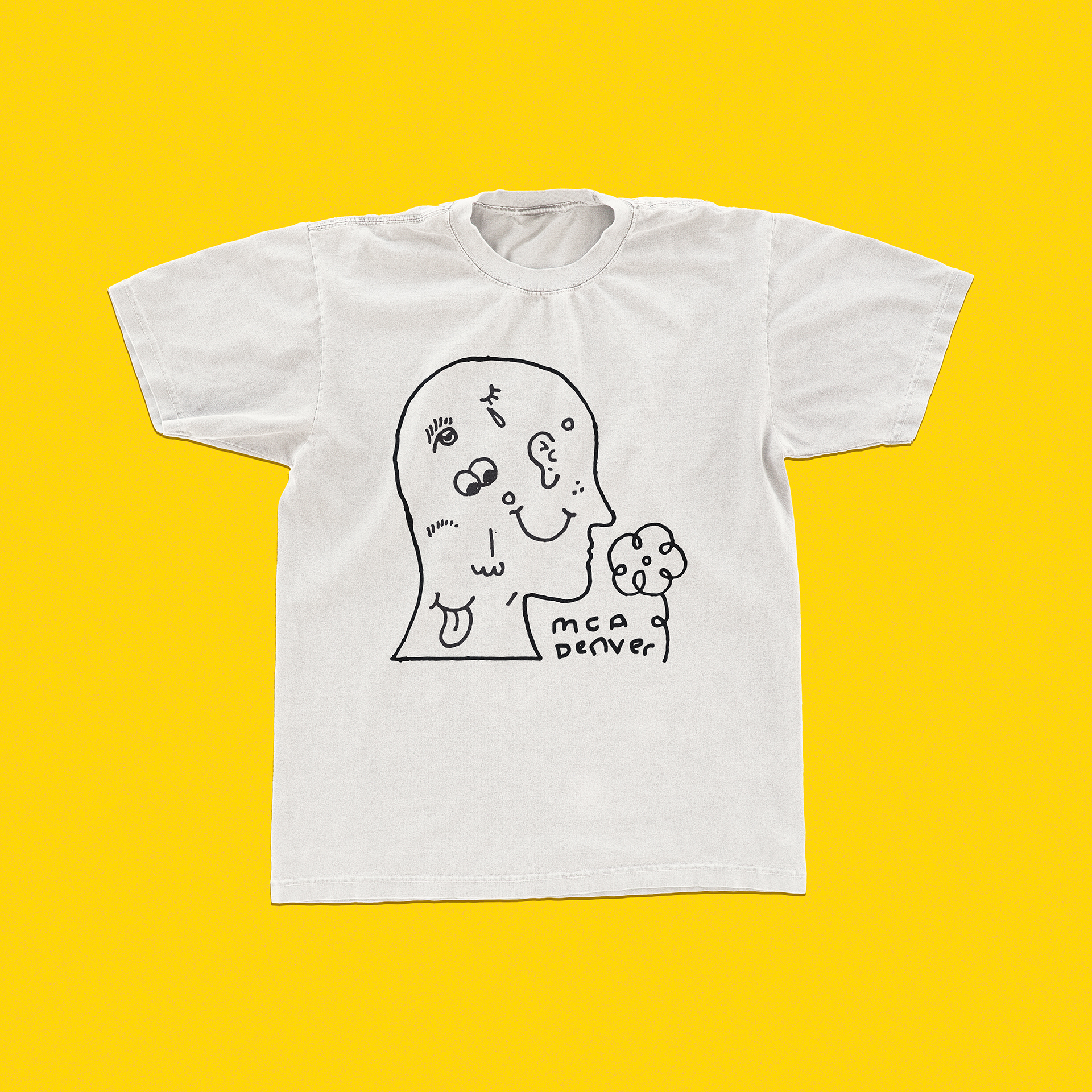 A white t-shirt on a yellow background with a black and white line drawing of the outline of a human profile. There is a small flower near the nose, which makes it look as though the figure on the shirt is smelling the flower. There is text between the flower and the head that reads “MCA Denver”. Inside the head are eyes, an ear, a smiley face, a mouth with a tongue sticking out, and a few other facial features.