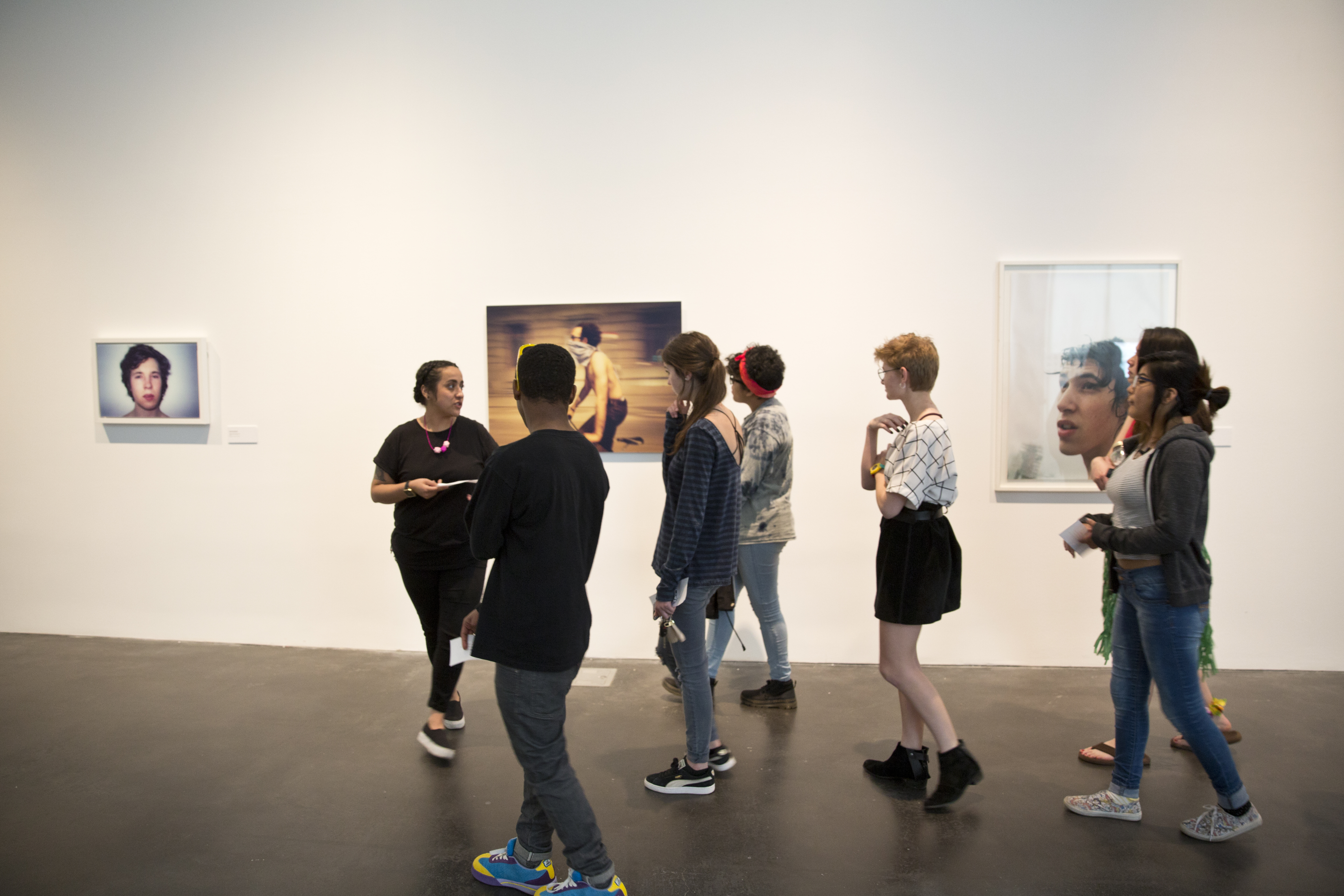 A group of teens are led by an adult in an art gallery. The walls hold photophraphs of young men. 