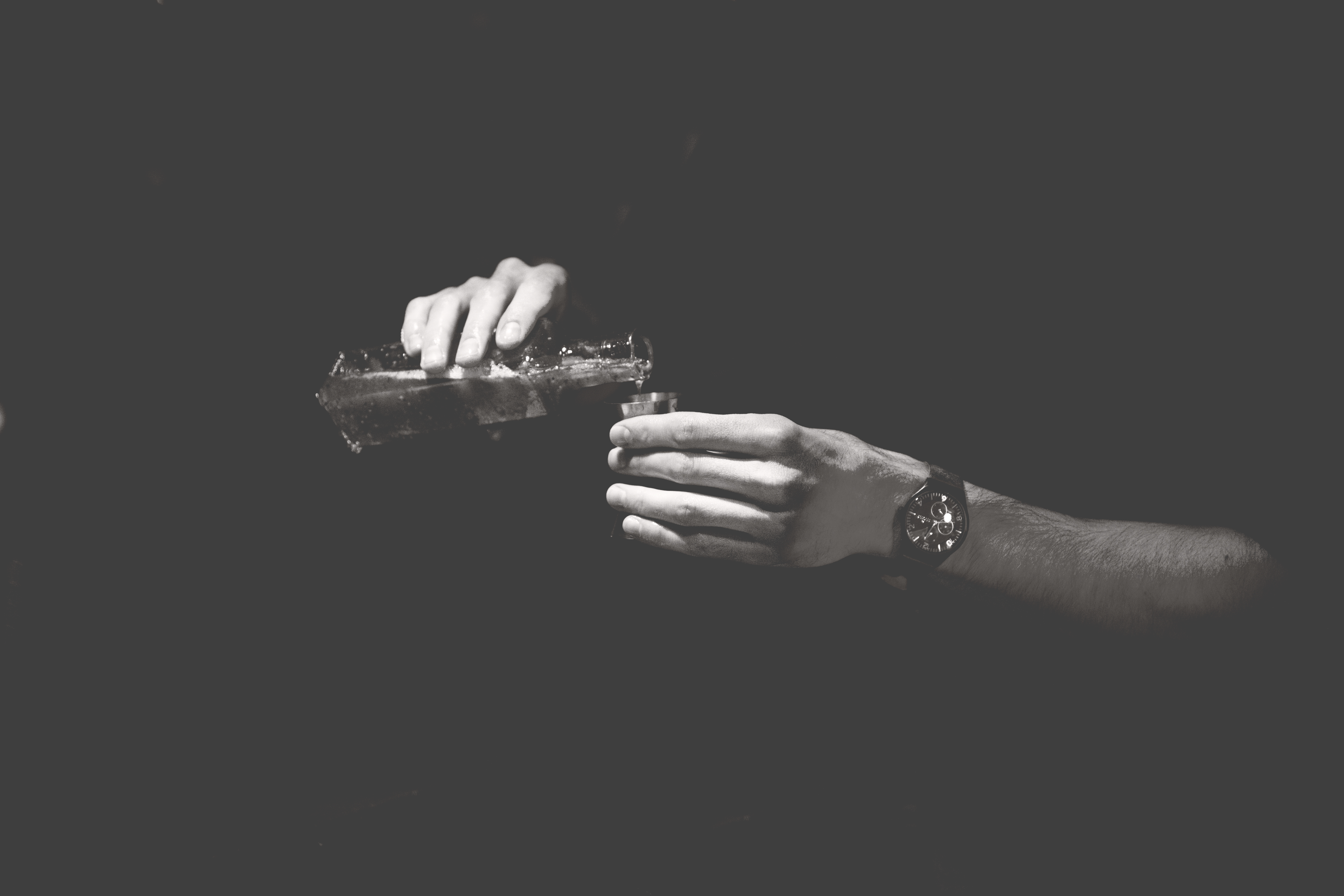 A black and white image of hands pouring a liquid into a measuring jigger. The body isn’t visible so it appears as if the hands are floating. 