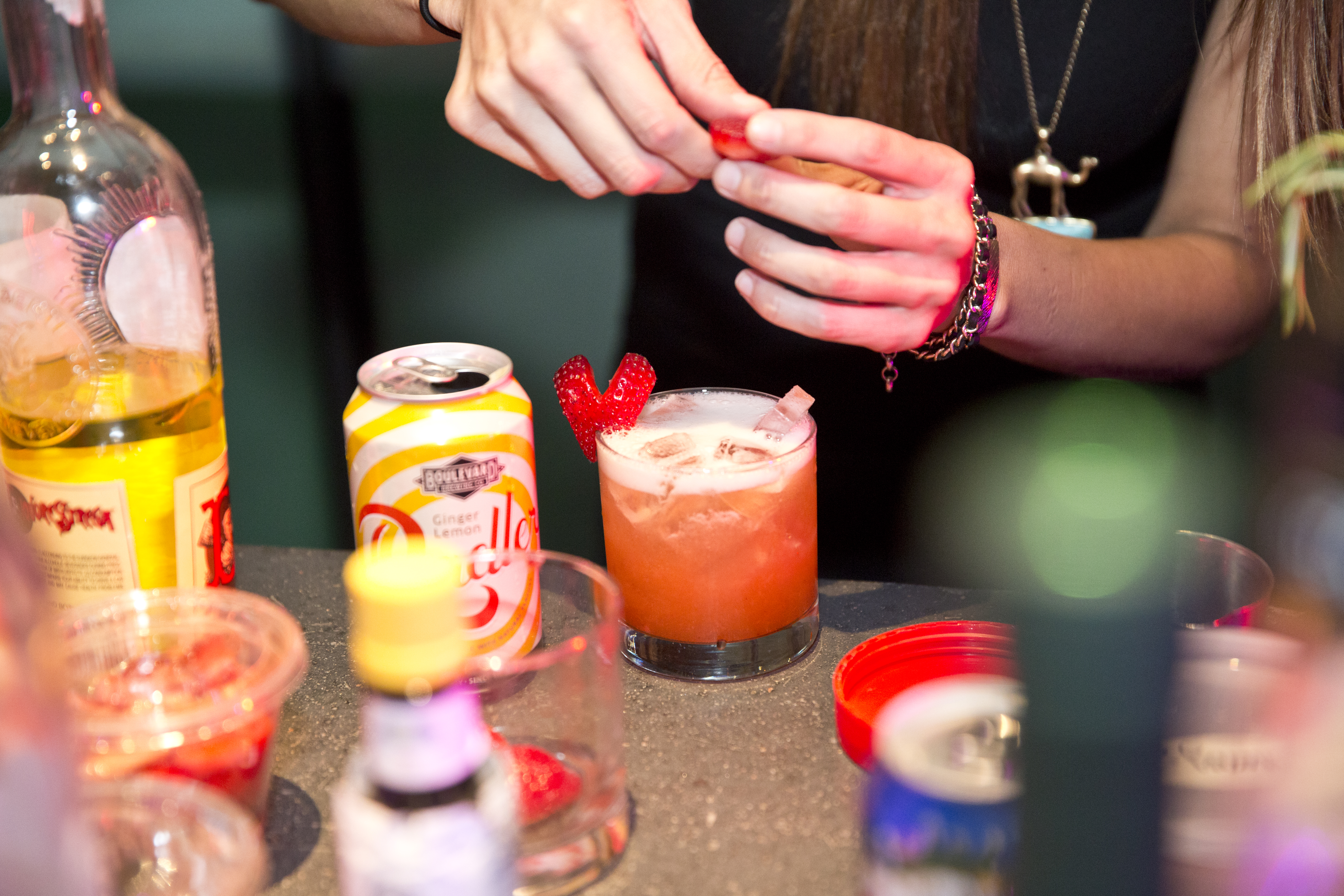 Hands prepare a blood orange colored cocktail on a cluttered bar. The drink is garnished with a strawberry in the shape of a heart.